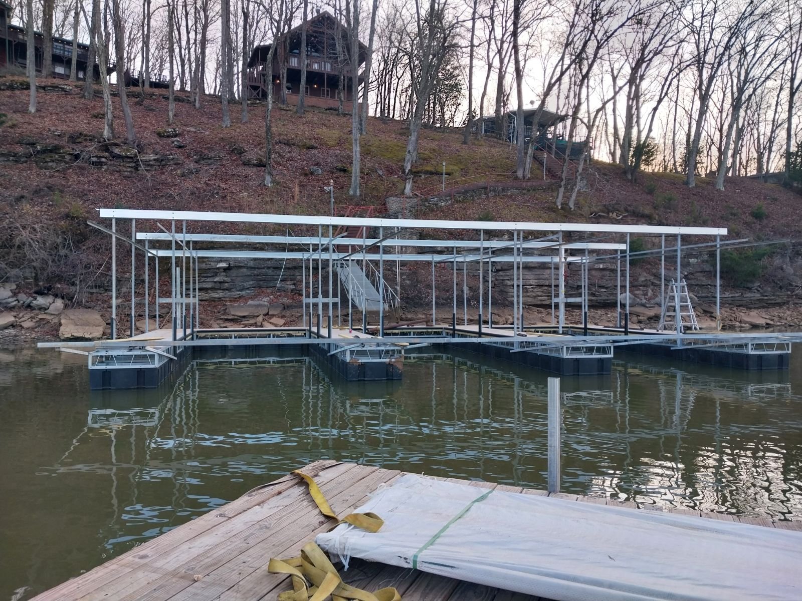 Underwater dock in the process of construction, with metal framework partially assembled, floating on a lake near a wooded hillside with bare trees and residential houses at the top.