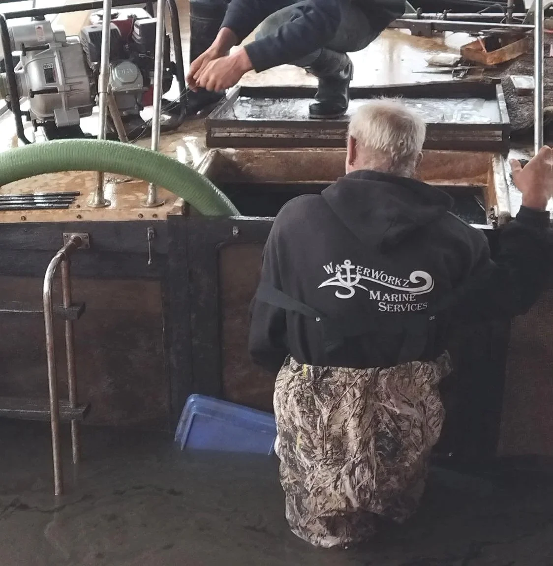 A person kneeling on the ground in camouflage pants and a black jacket with marine services logo, working on a boat engine with another person standing on the boat. The boat’s surface and engine are visible.