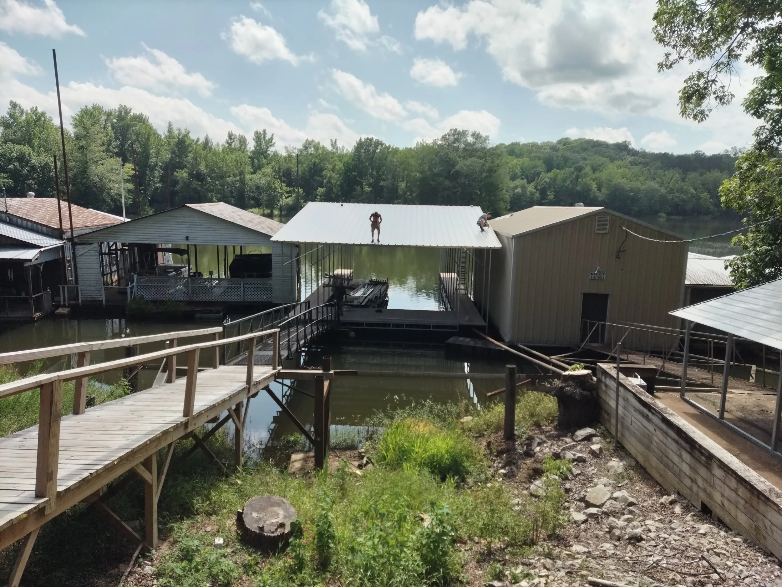 A lakeside scene with docks and small buildings, including a house with a white metal roof, trees in the background, and a partly cloudy sky.