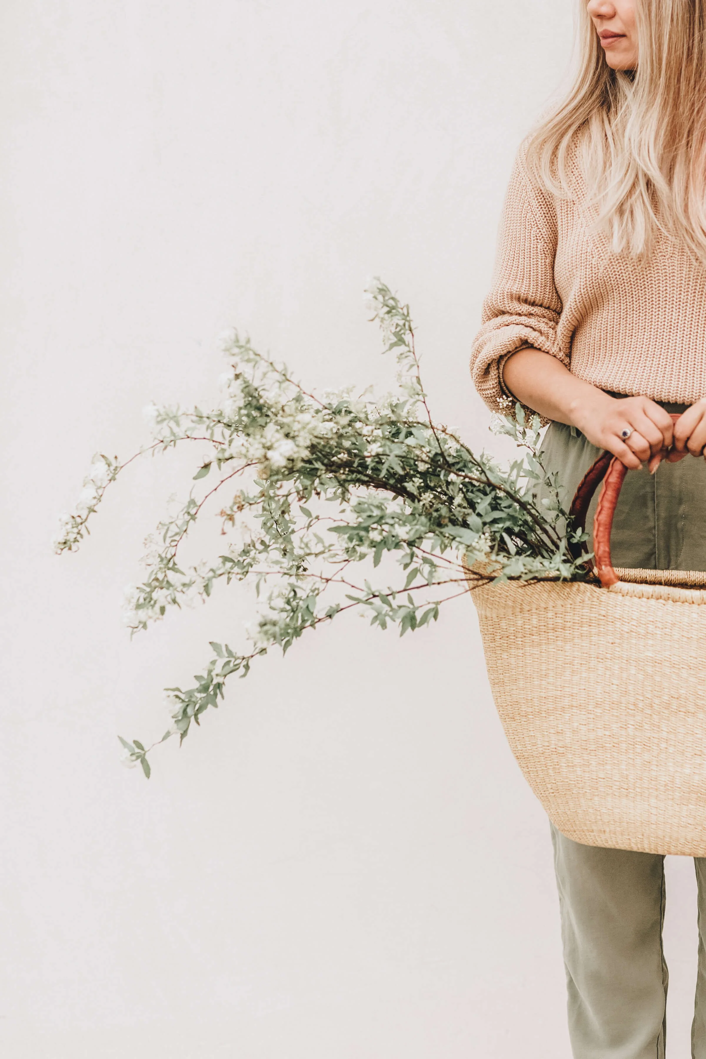 A woman holding a woven basket with green foliage and white flowers against a plain white background.