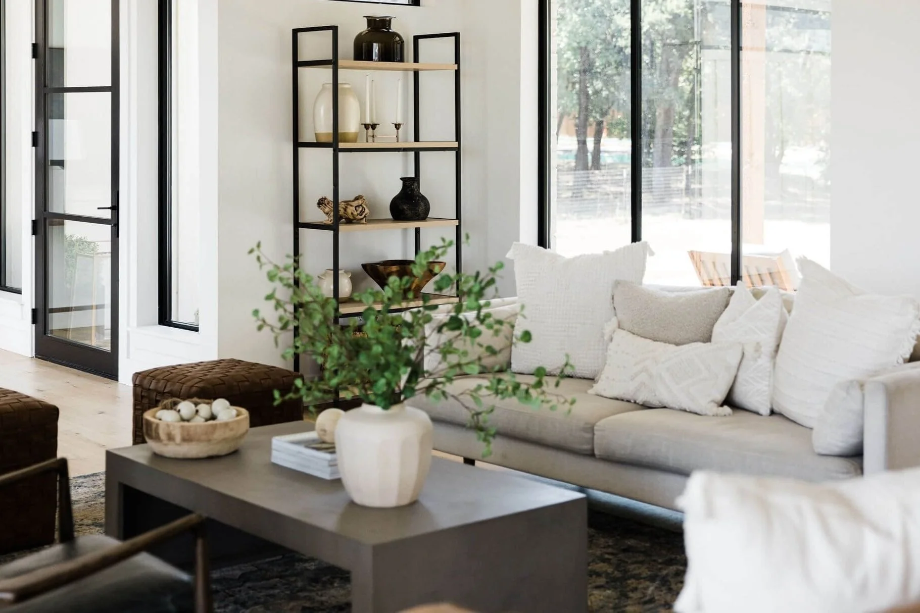 Integrative Nutrition office with large windows, beige sofa with white pillows, black metal and wood shelving unit with decorative items, green plant in a white vase on a gray coffee table, and sunlight streaming through the windows.