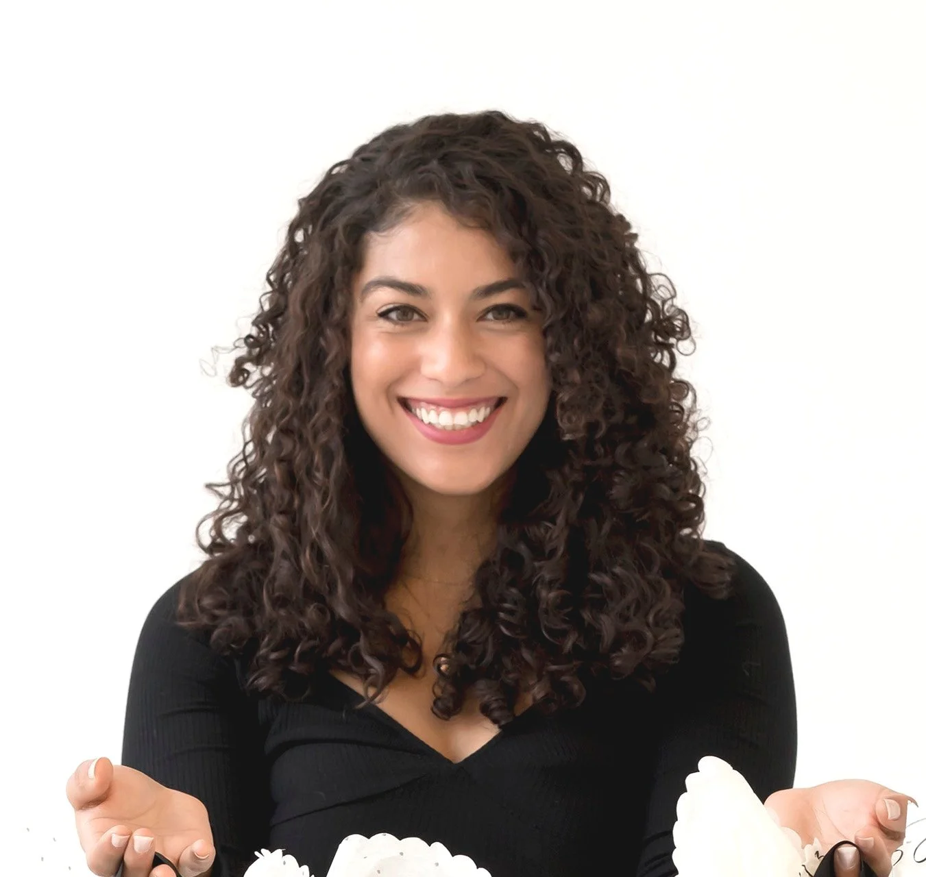 Smiling woman with curly brown hair wearing a black top, holding white flowers against a white background.