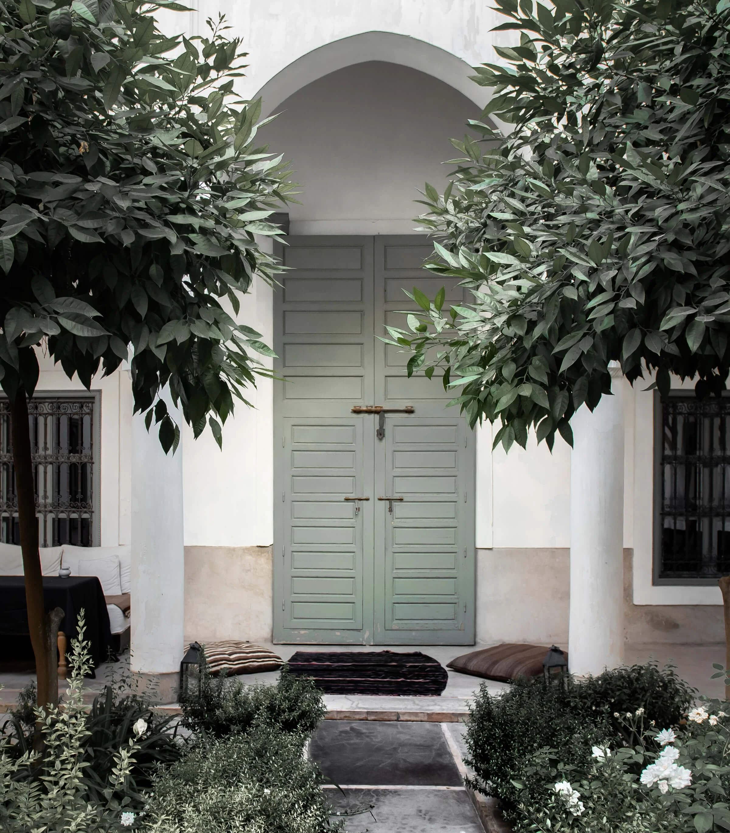 A front door of a house painted in light green with wooden shutters, framed by white walls and surrounded by garden greenery, including trees, bushes, and flowering plants.