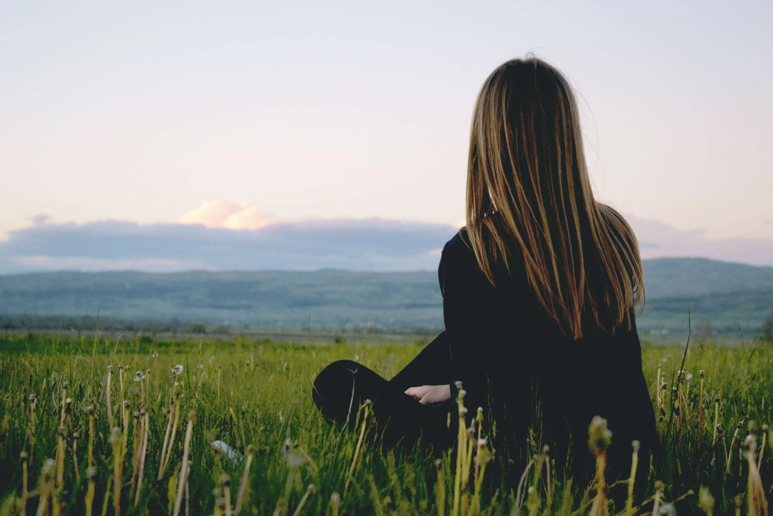 A woman with long hair sitting in a grassy field facing away, gazing at distant mountains and a cloudy sky at sunset.