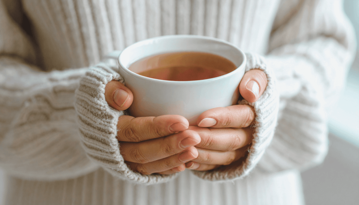 Person holding a white mug of tea while wearing cozy fingerless gloves and a beige sweater.