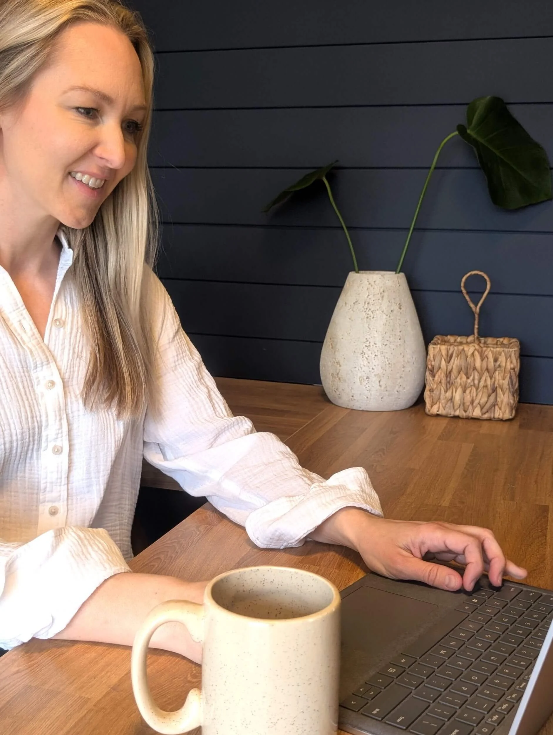 Lindsay Christen (Integrative Nutritionist, Menopause Specialist) sitting at her desk, smiling while using a laptop. There is a beige ceramic mug and a potted plant with large green leaves in a white vase behind her on a dark blue wall.