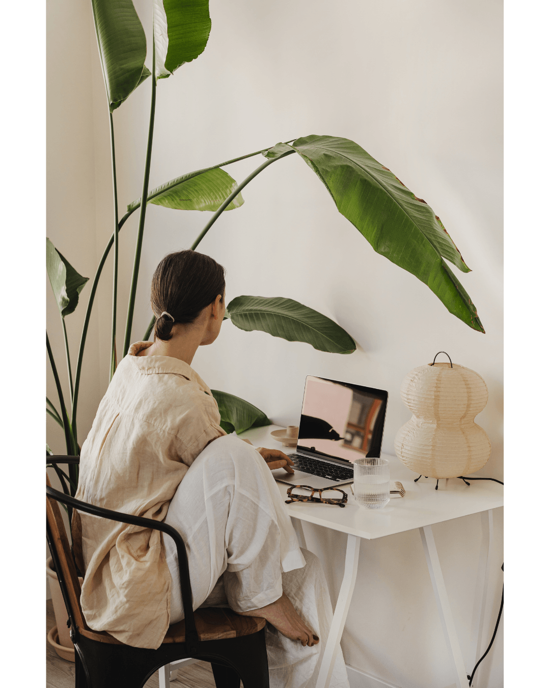 A woman with brown hair in a bun, wearing a beige shirt, sitting at a white desk with a laptop, large green plant, glass of water, eyeglasses, and a paper lantern lamp.