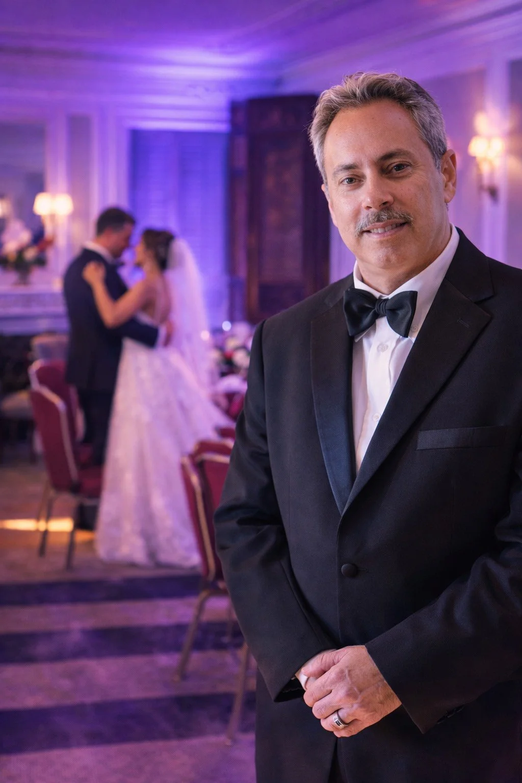 A man in a black tuxedo with a bow tie smiling at the camera at a formal wedding reception, with a bride and groom dancing in the background.