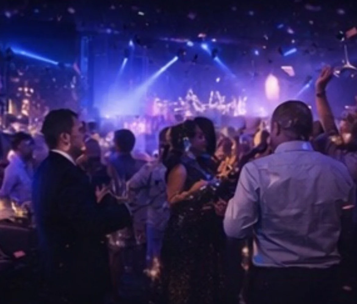 People dancing and socializing in a nightclub with blue and purple lighting, a live DJ or band performing on stage in the background.
