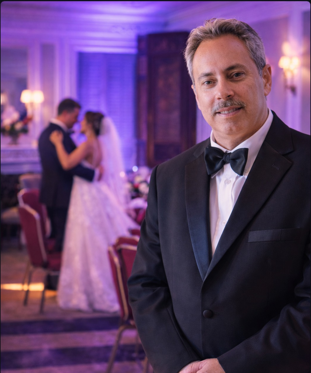 A man in a tuxedo with a bow tie smiling in the foreground at a wedding reception. In the background, a bride and groom are dancing closely.
