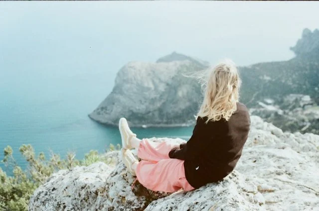 A blonde woman with her back at the camera sitting on a rock looking into the distance over the ocean.