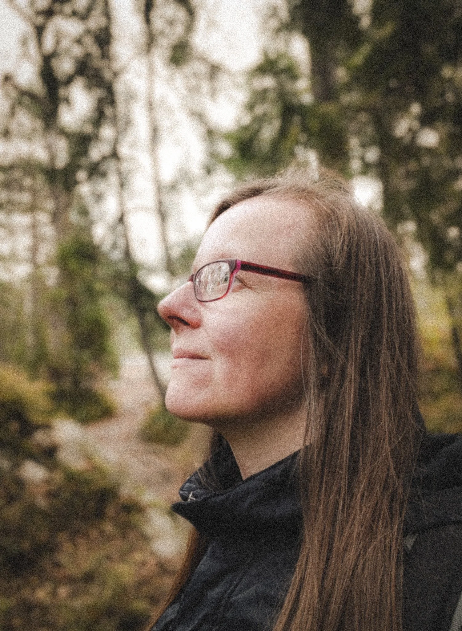 A woman with long brown hair, wearing glasses and a black jacket, smiling outdoors near trees with green and yellow leaves.