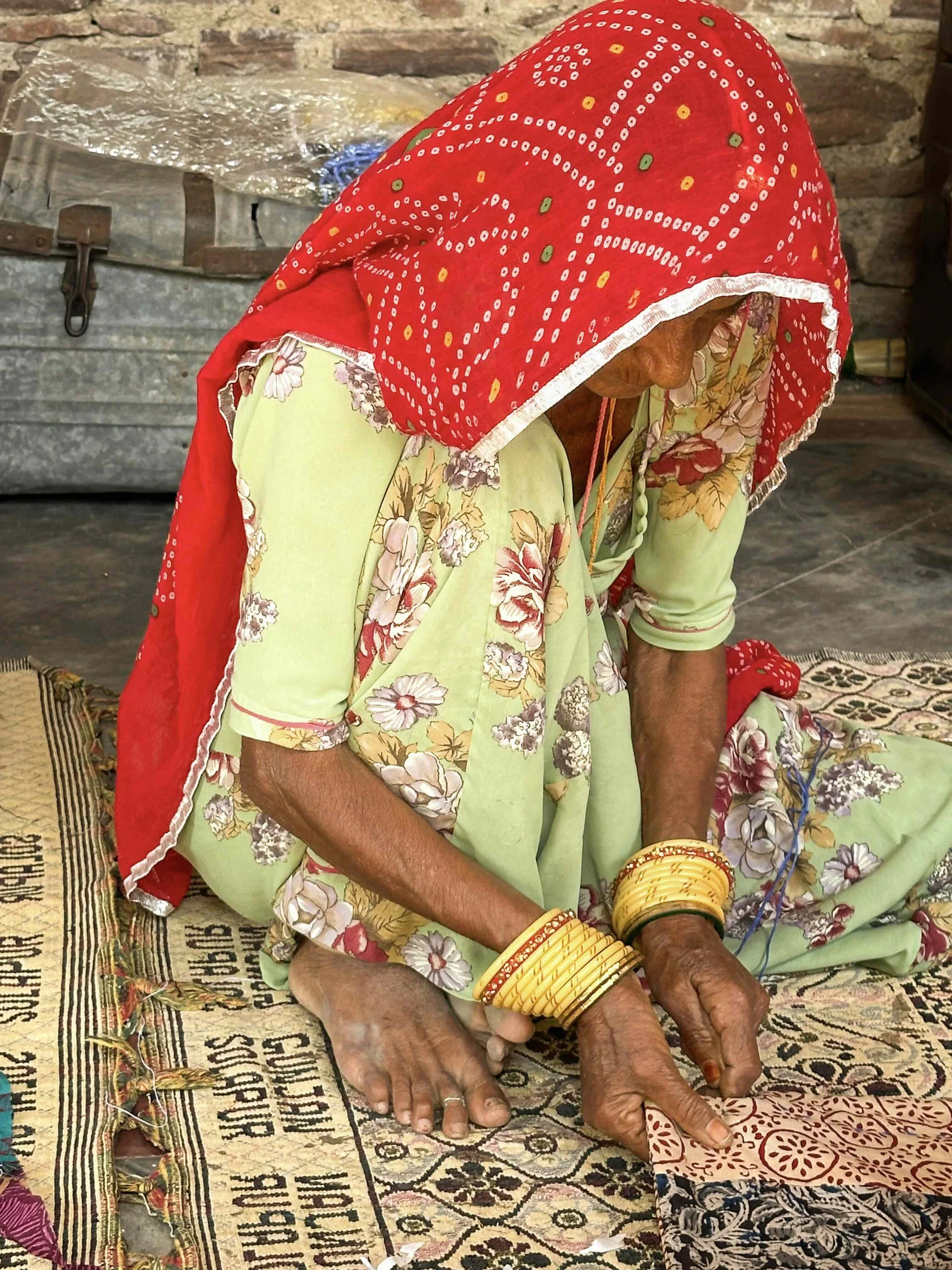An older Rajasthani woman seated and embroidering at Sunder Rang, Chandelao, wearing a red bandhani odhni drawn forward over her face and gold bangles, her hands working the cloth.