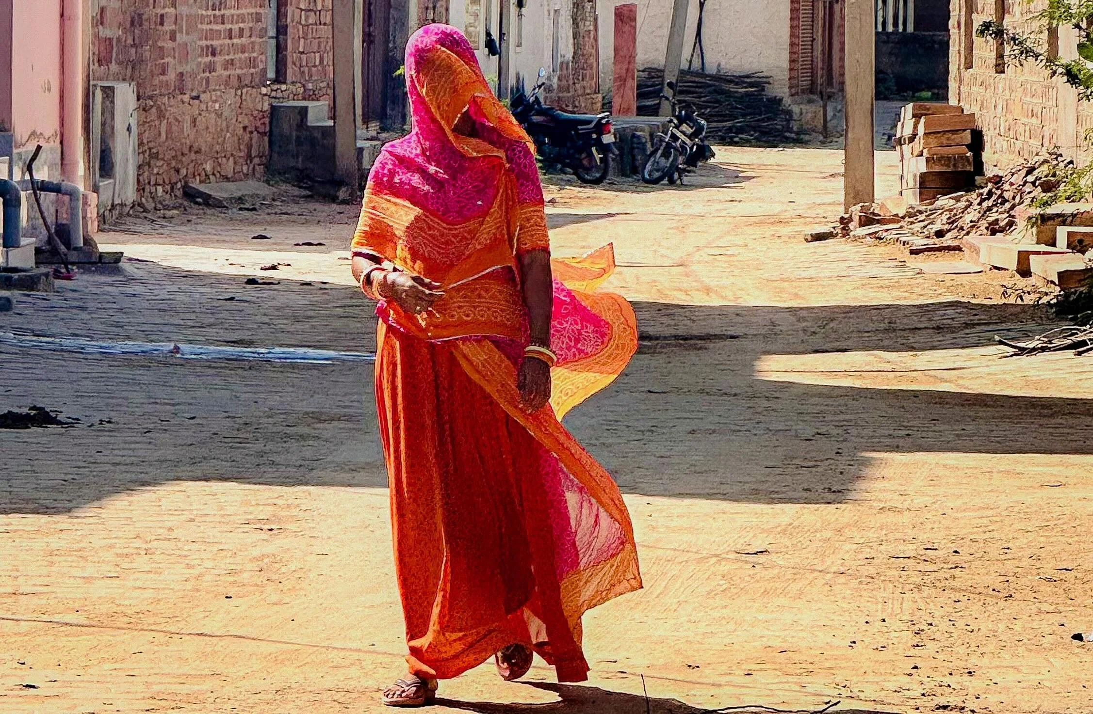 A Rajasthani woman walking across a sunlit village courtyard, dressed in orange and pink ghagra and odhni, her pallu drawn forward over her face.