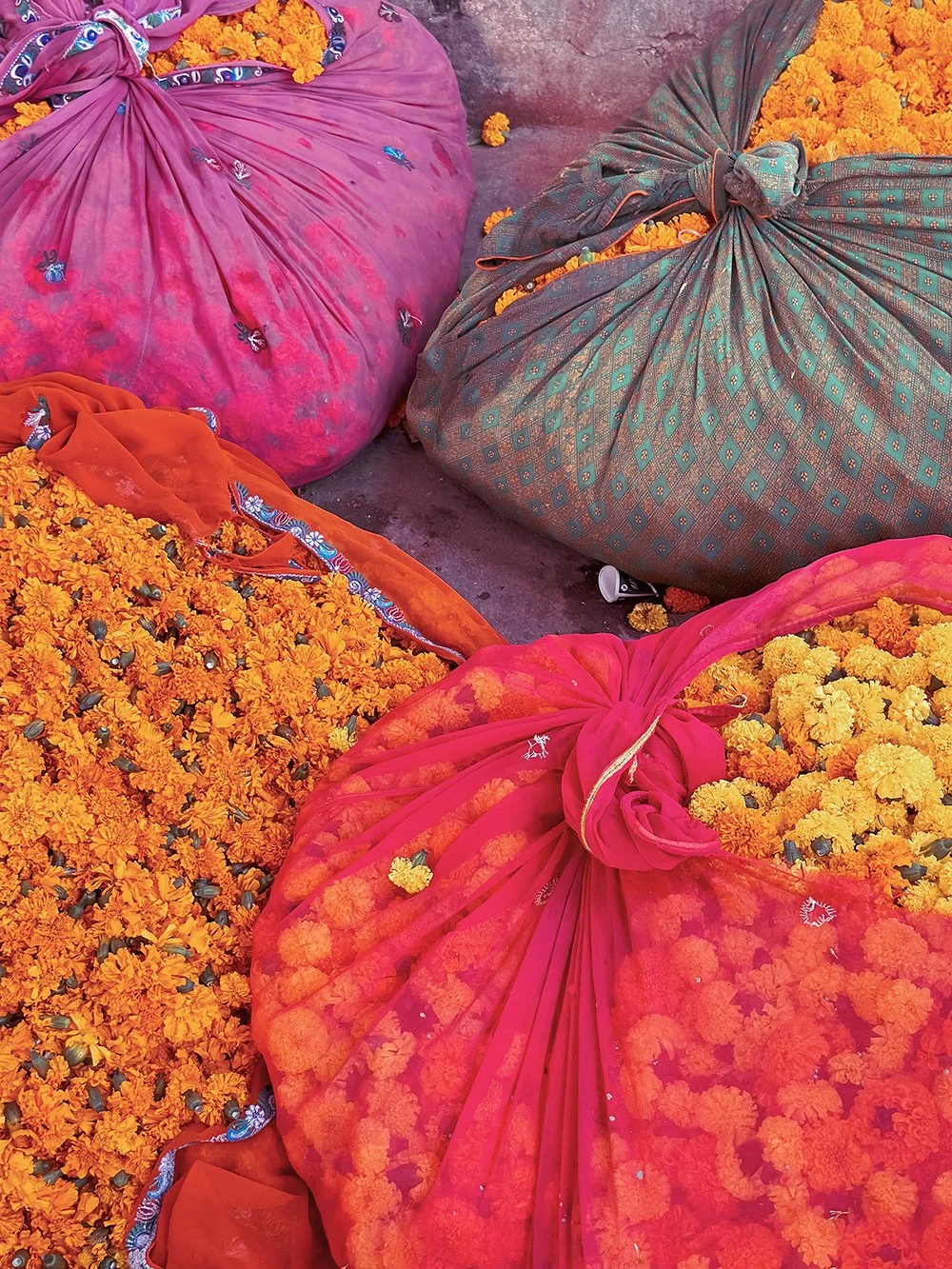 Colorful bunches of orange and yellow marigold flowers next to fabric and large pink and green sacks tied at the top, likely used for carrying or storing flowers. Bright, festive scene possibly from a flower market or celebration.