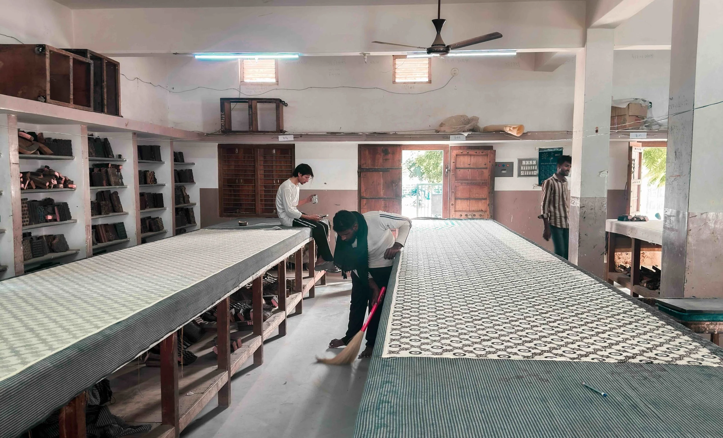 The interior of Khatri Sufiyan Ismail’s ajrakh workshop in Ajrakhpur — long printing tables, shelves lined with wooden blocks, and two printers at work in a light-filled, ordered space.
