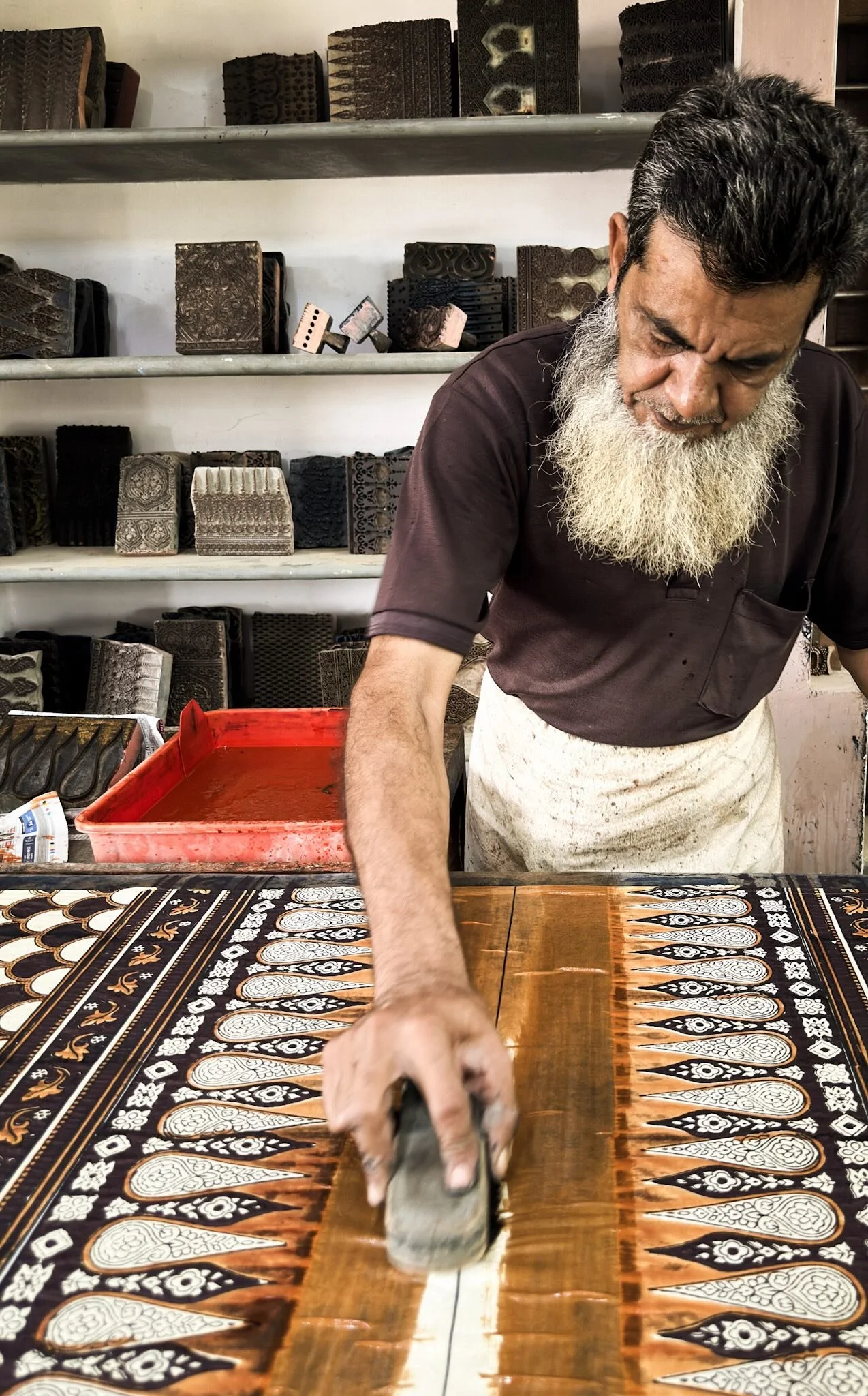 Master ajrakh printer Rafiq Bhai Khatri pressing a printing block onto fabric at the printing table at Sufiyan Ismail Khatri workshop Ajrakhpur Kutch Gujarat shelves of carved wooden blocks behind him