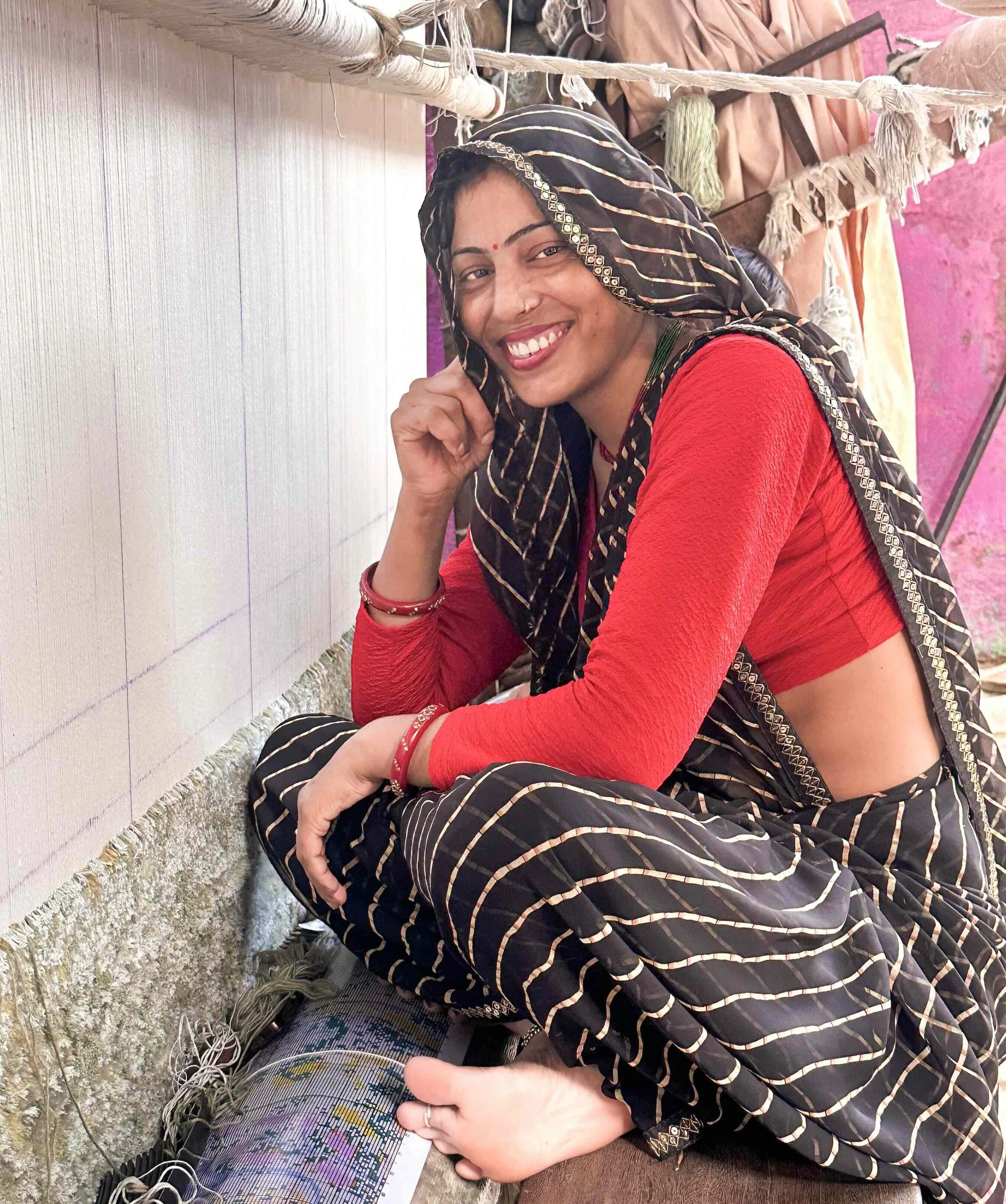 A Rajasthani weaver seated at her loom in a village outside Jaipur, smiling, wearing a black and white leheriya odhni with silver border and red choli, weaving for Jaipur Rugs.