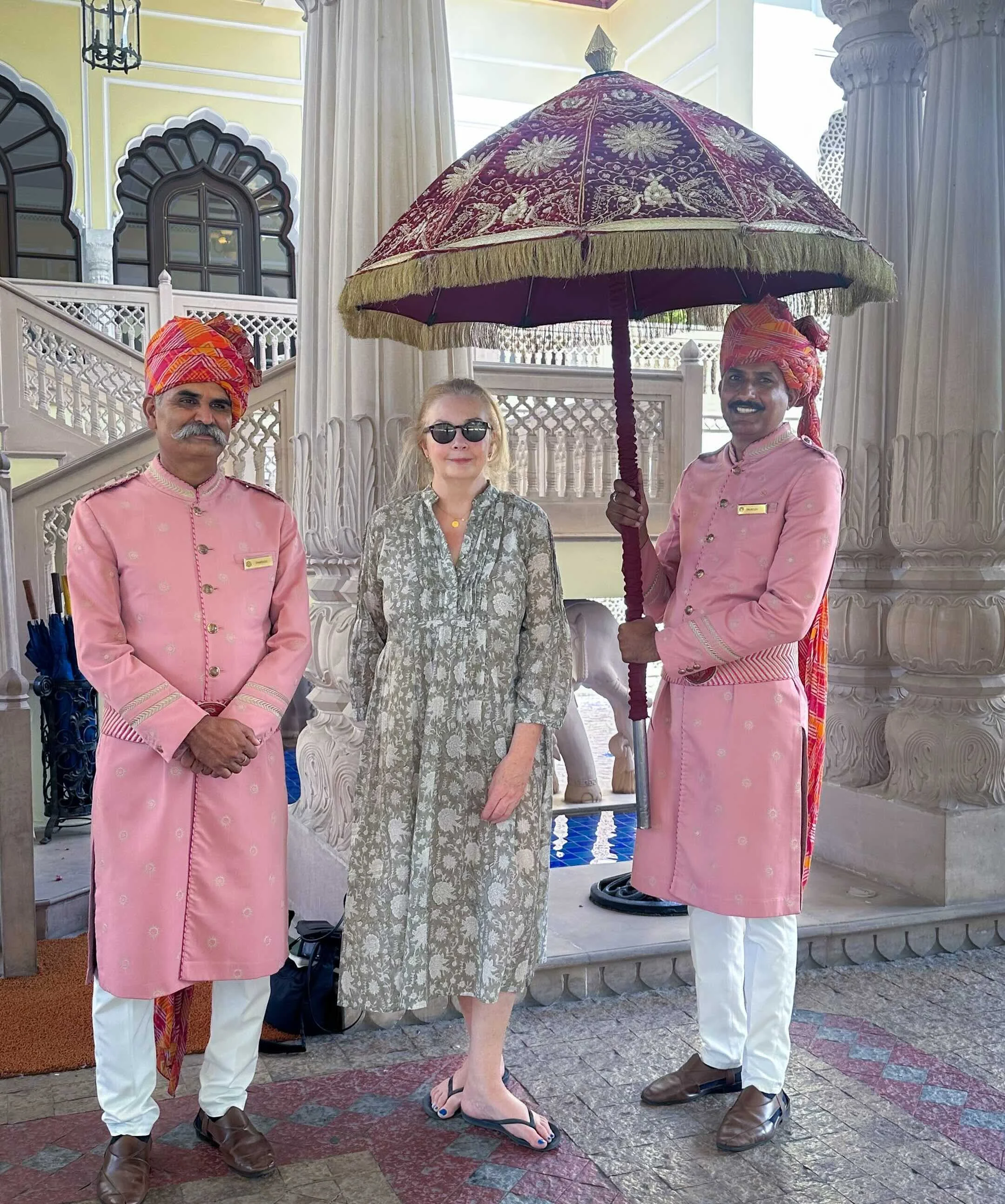 Bernadette O'Farrell standing beneath a traditional Indian ceremonial umbrella with two staff members in pink livery at Rambagh Palace, Jaipur