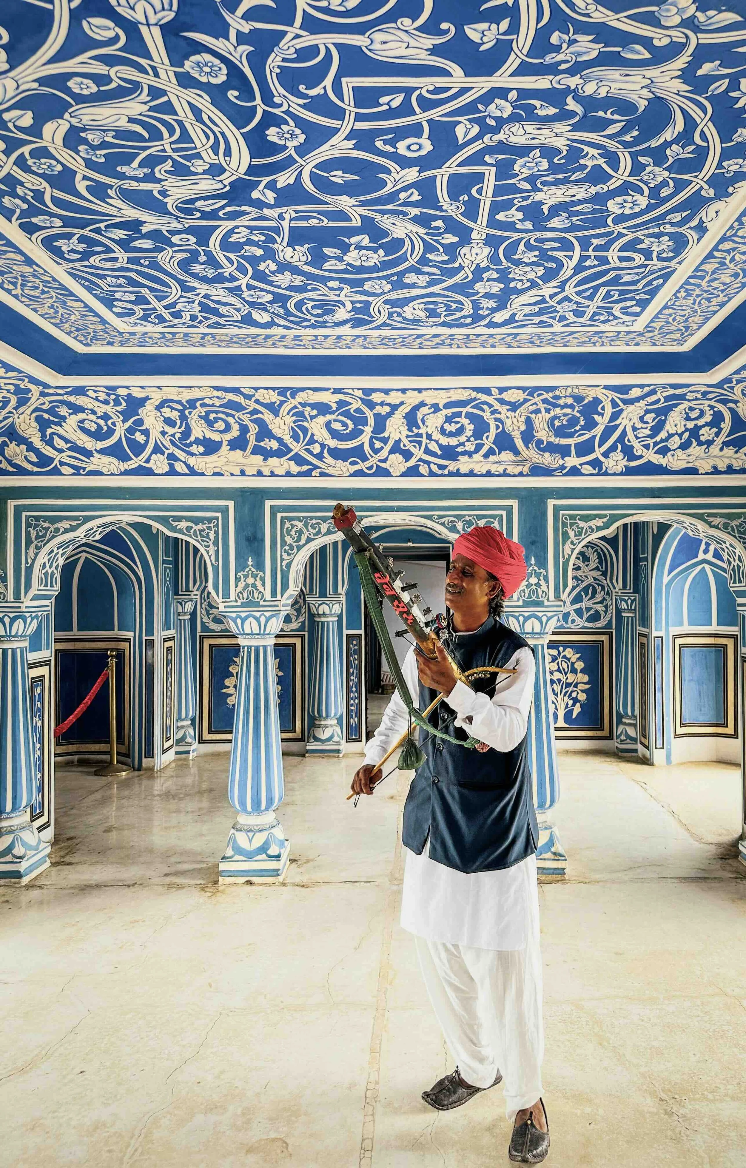 A musician in a red turban holding a stringed instrument stands in the Sukh Niwas, the hand-painted blue and white room of the Chandra Mahal at City Palace, Jaipur, the ornate painted ceiling above him.​​​​​​​​​​​​​​​​