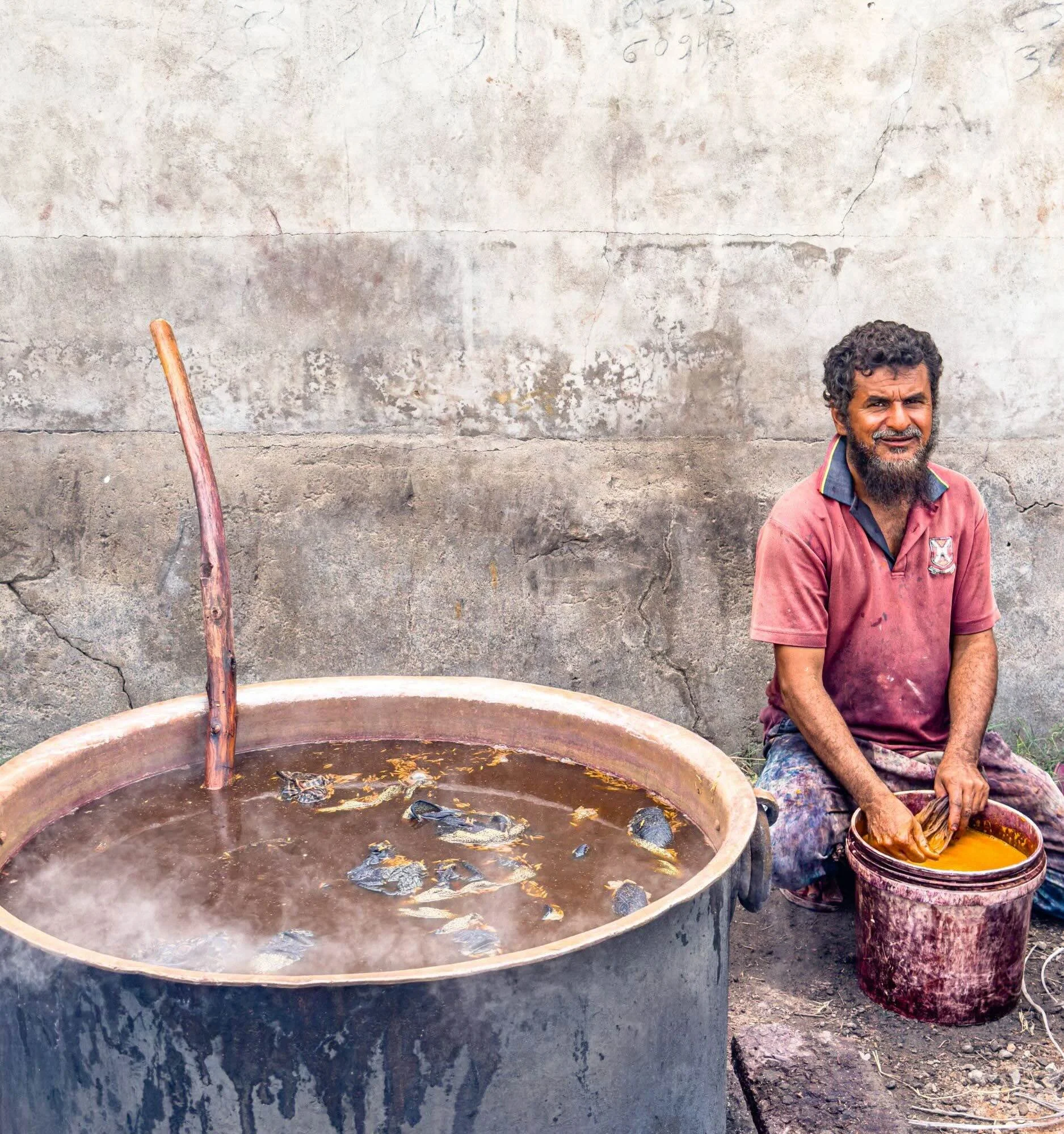 An ajrakh dyer sitting beside a large outdoor dyeing vat in Ajrakhpur, Kutch, with a bowl of dye beside him.