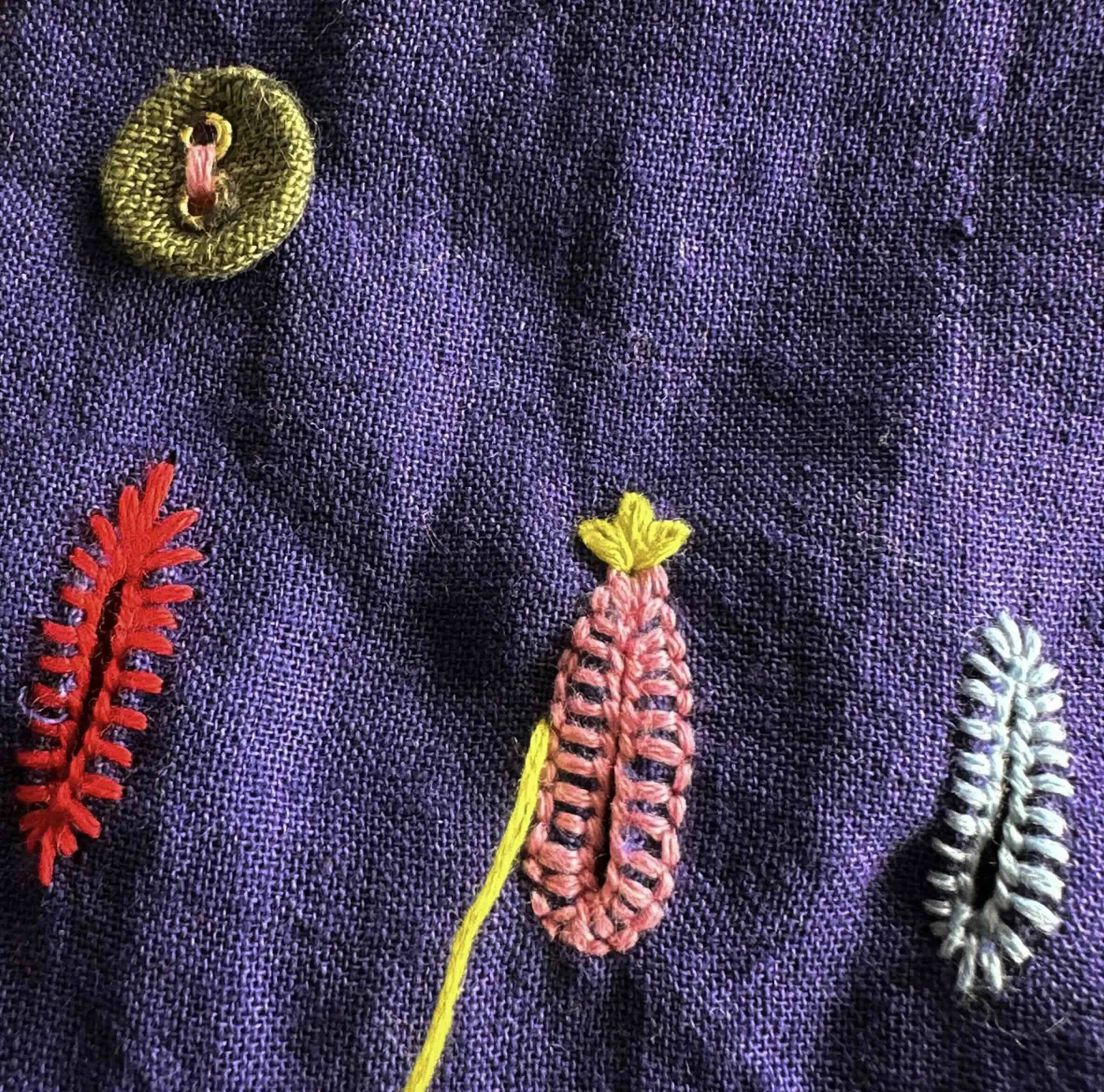 Close-up of hand embroidery on indigo fabric at Artisav guesthouse, Jaipur, showing decorative buttonhole stitching and embroidered motifs in red, pink, yellow and white thread.​​​​​​​​​​​​​​​​