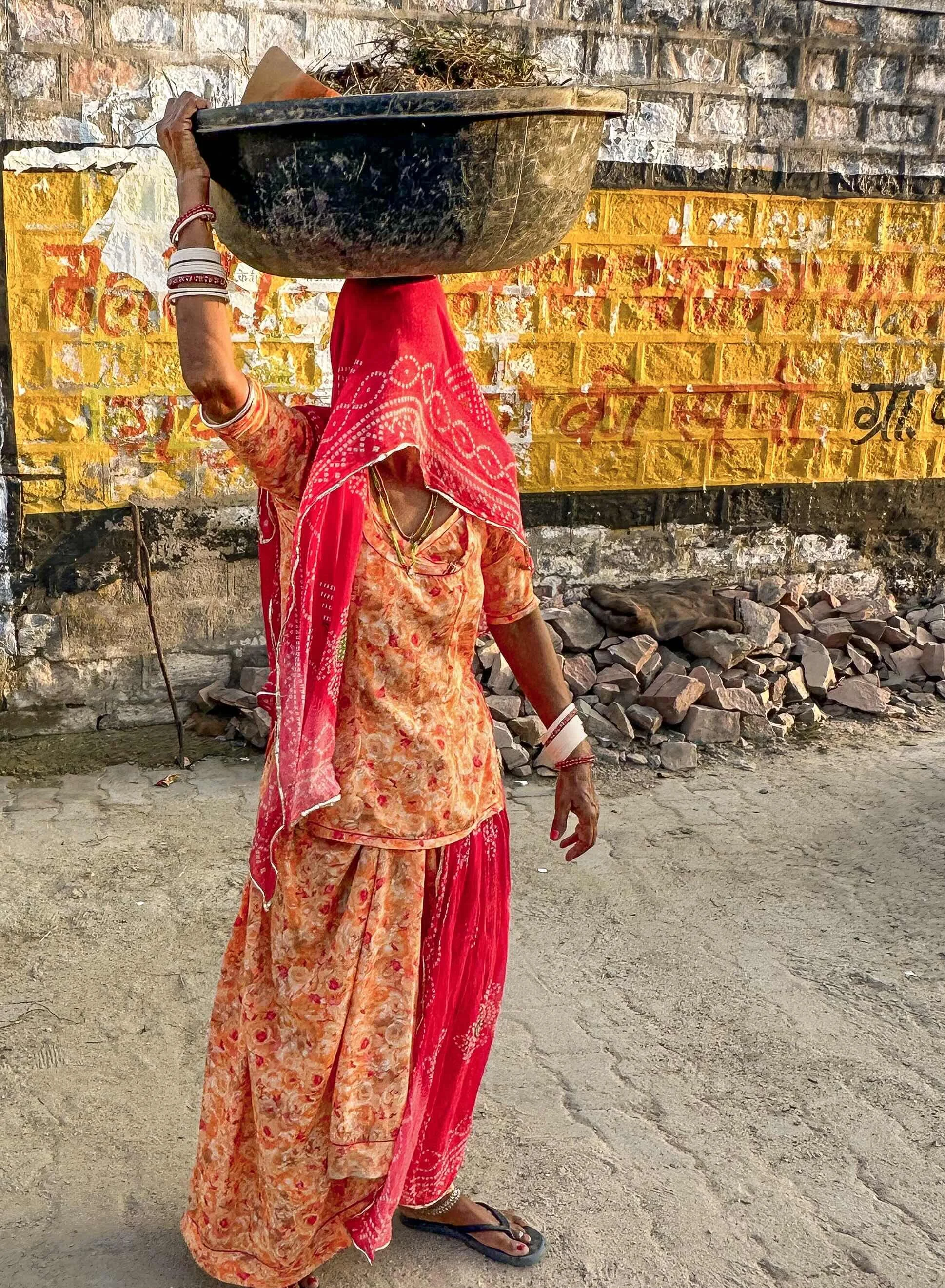 A Rajasthani woman balancing a large metal bowl on her head, wearing a pink and red bandhani odhni and ghagra with white bangles stacked to the elbow, walking past a painted village wall.​​​​​​​​​​​​​​​​