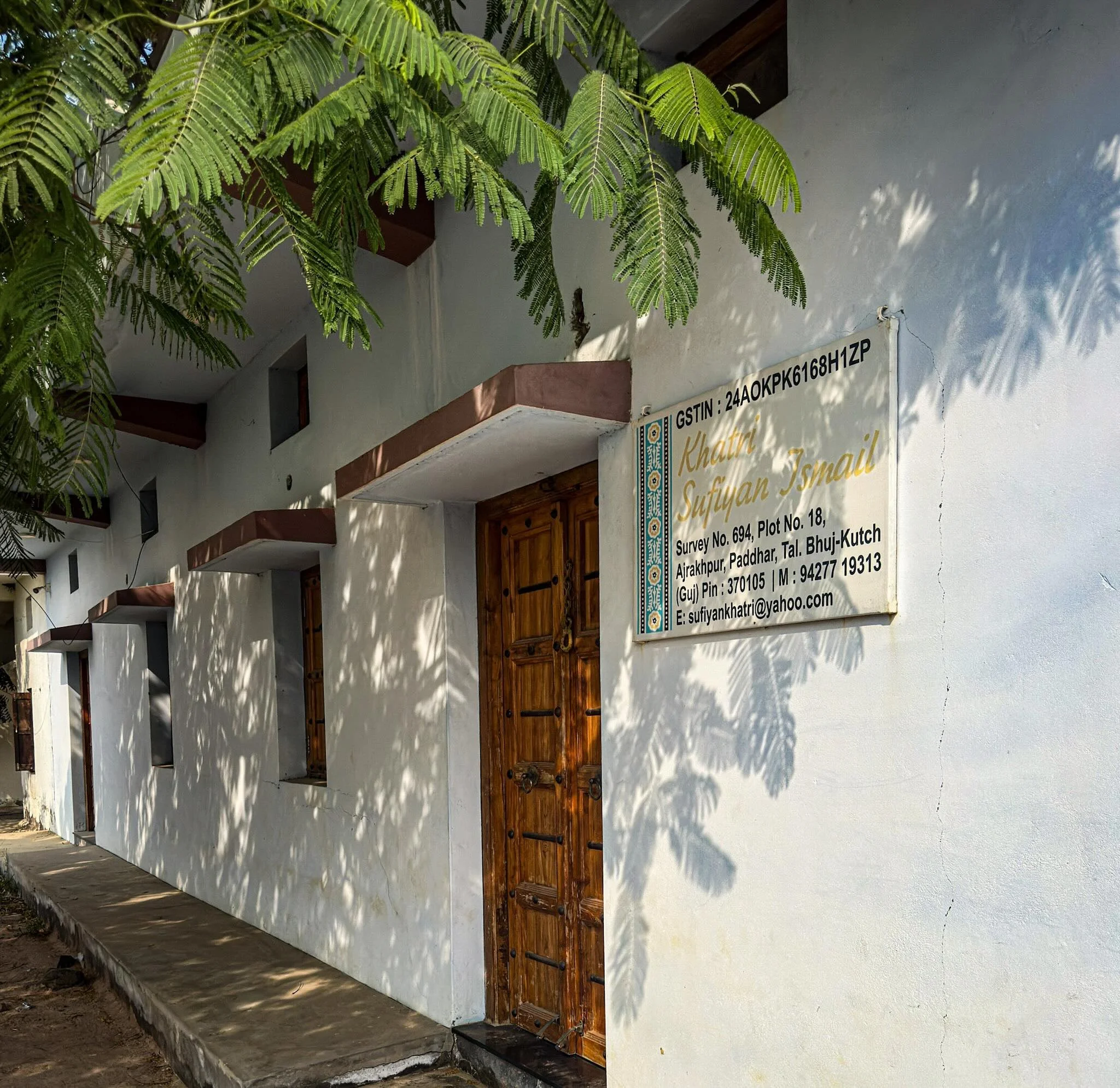 Exterior of Khatri Sufiyan Ismail ajrakh workshop, white building with wooden door and name sign, dappled light through trees, Ajrakhpur, Kutch, Gujarat