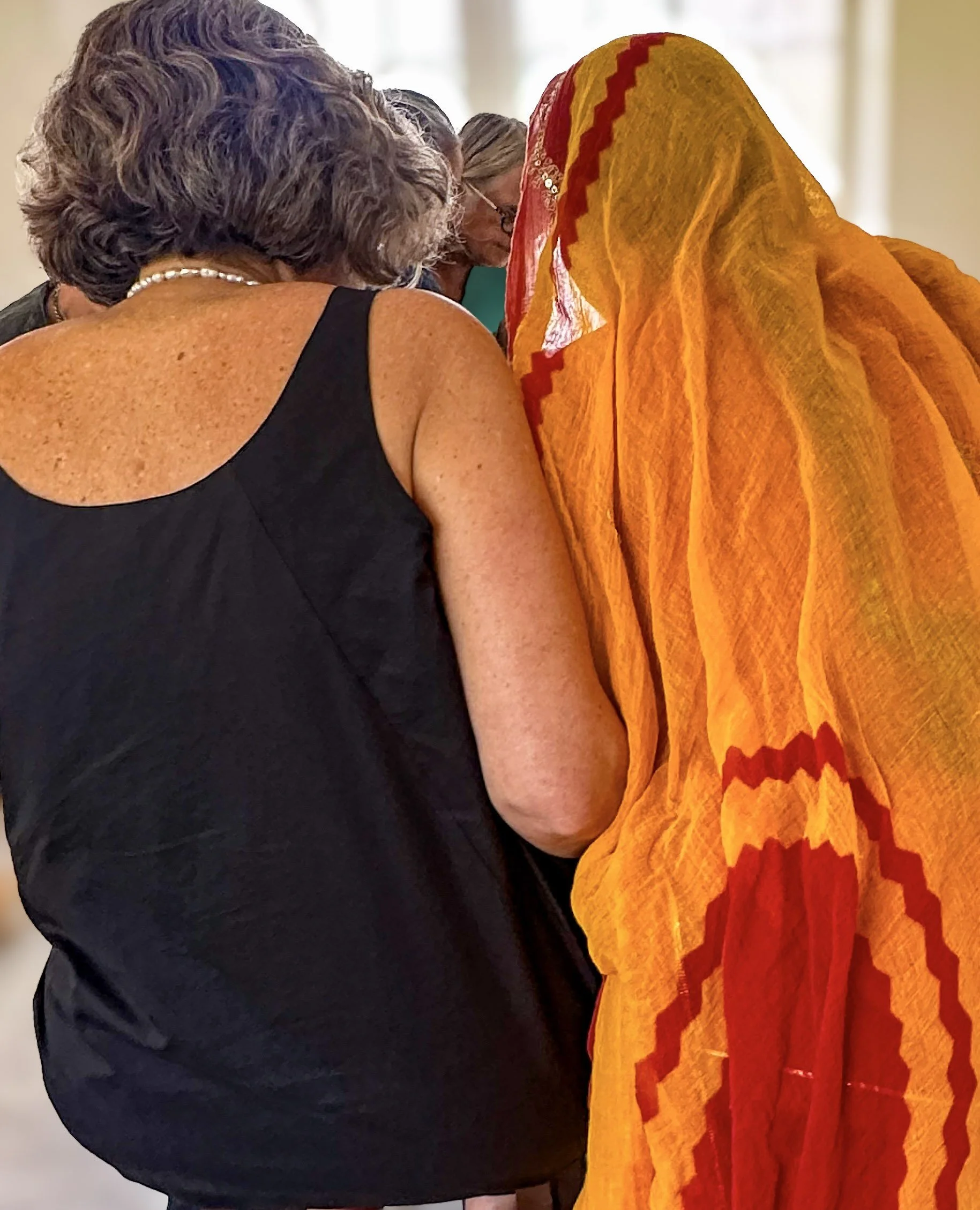Two women seen from behind at a gudri quilting workshop at Nila House, Jaipur, one in a black vest and one in a saffron and red odhni, heads close together as they examine the work.​​​​​​​​​​​​​​​​