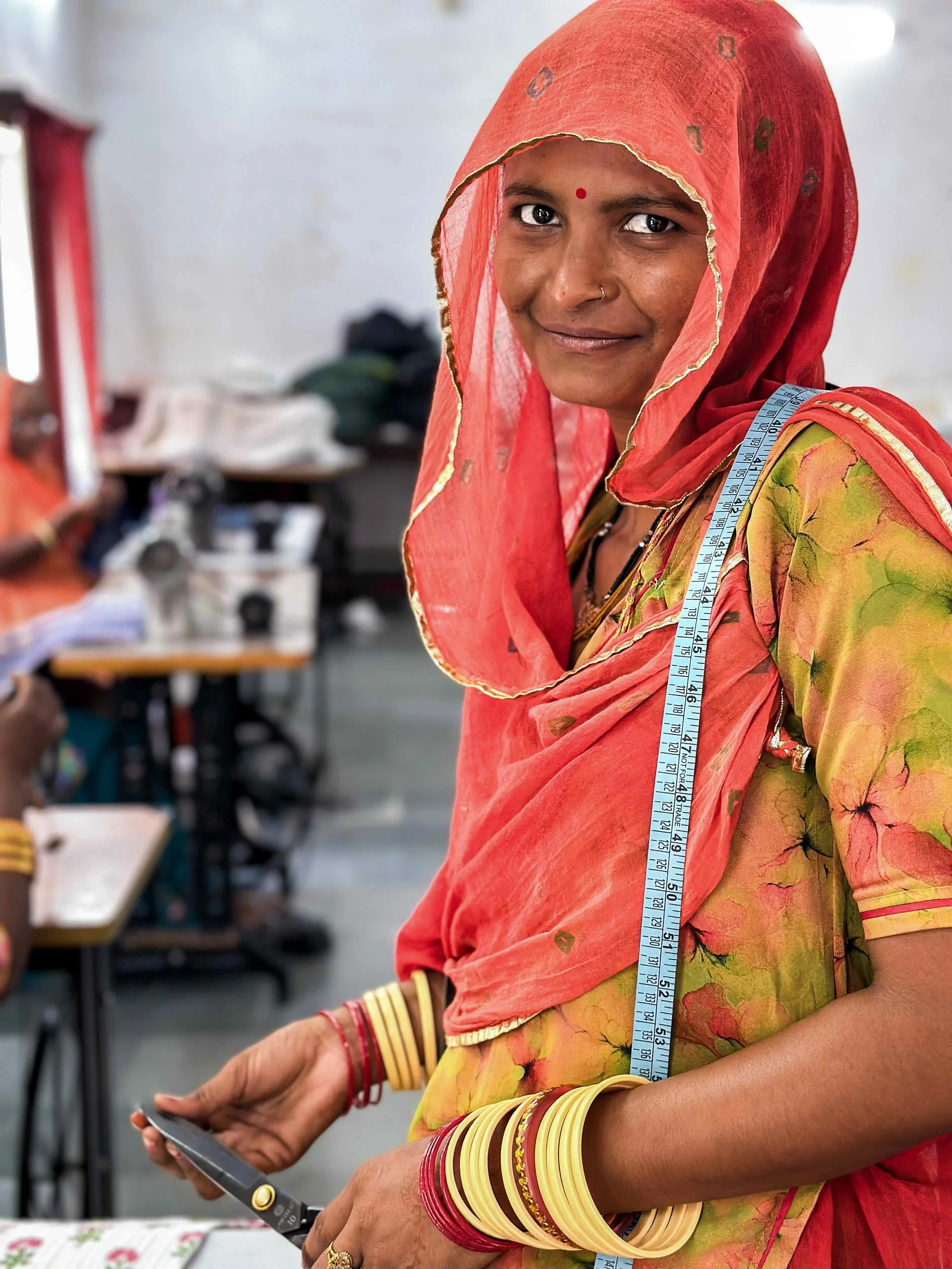 Portrait of a Rajasthani woman at Sunder Rang, Chandelao, wearing a red mulmul cotton odhni and yellow floral choli, a tailor’s measuring tape over her shoulder, looking directly at the camera.​​​​​​​​​​​​​​​​