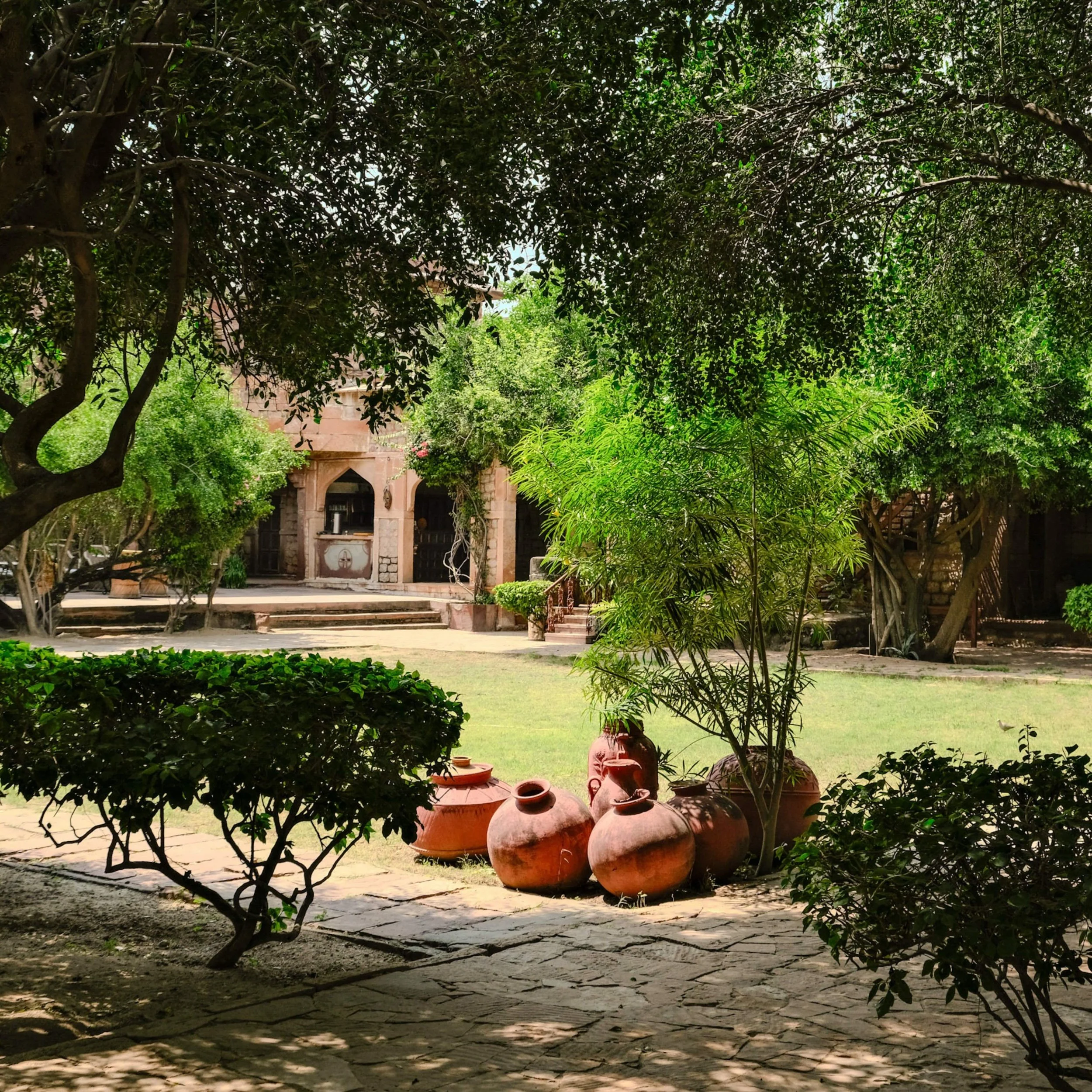 The walled courtyard garden of Chandelao Garh heritage property, Rajasthan — clusters of large terracotta water pots arranged on stone paving, surrounded by mature trees and clipped hedges.