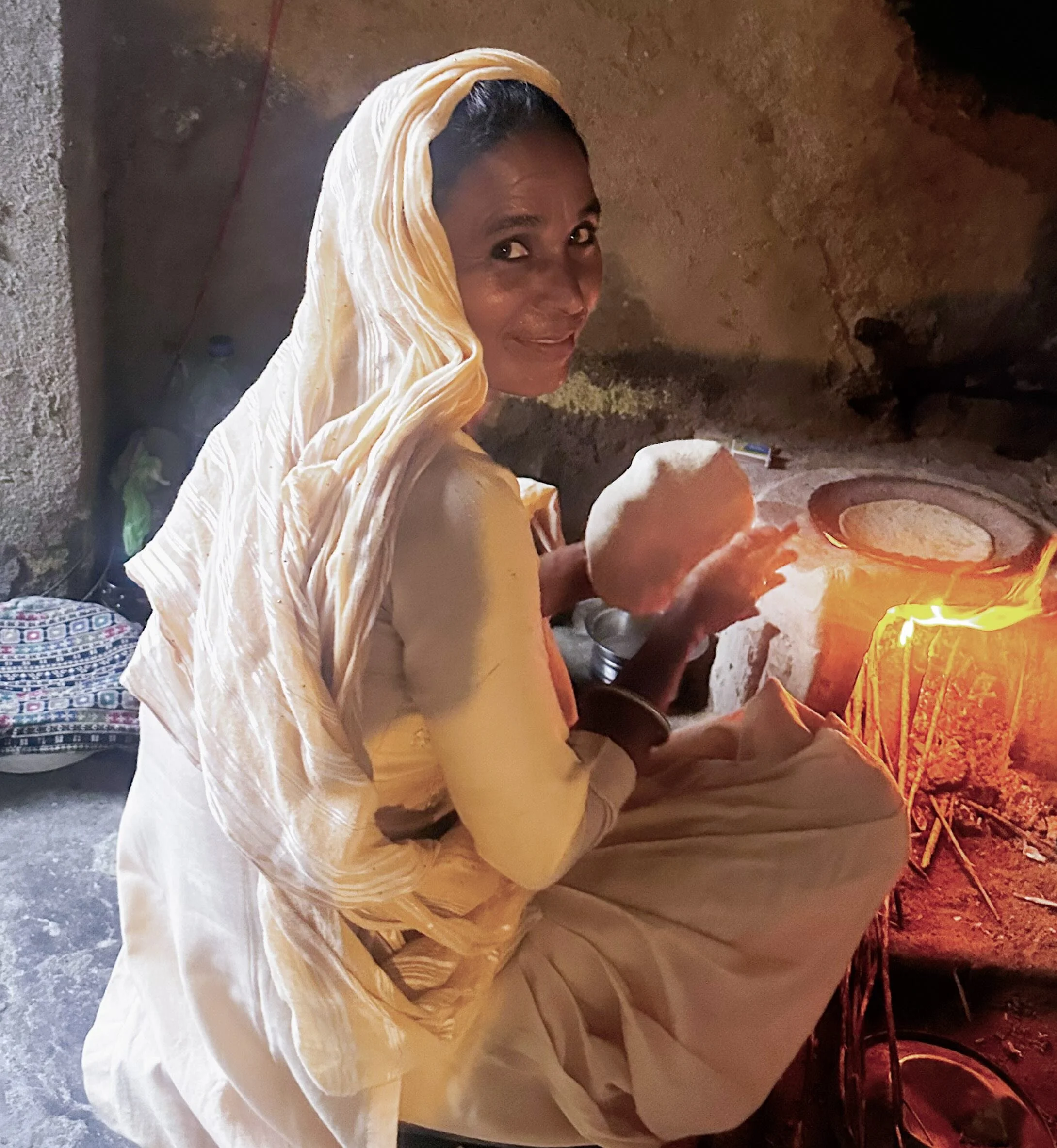 A Rajasthani woman seated by an open fire making chapatis in a village kitchen near Kesroli, wearing a pale cotton odhni, looking directly at the camera.​​​​​​​​​​​​​​​​