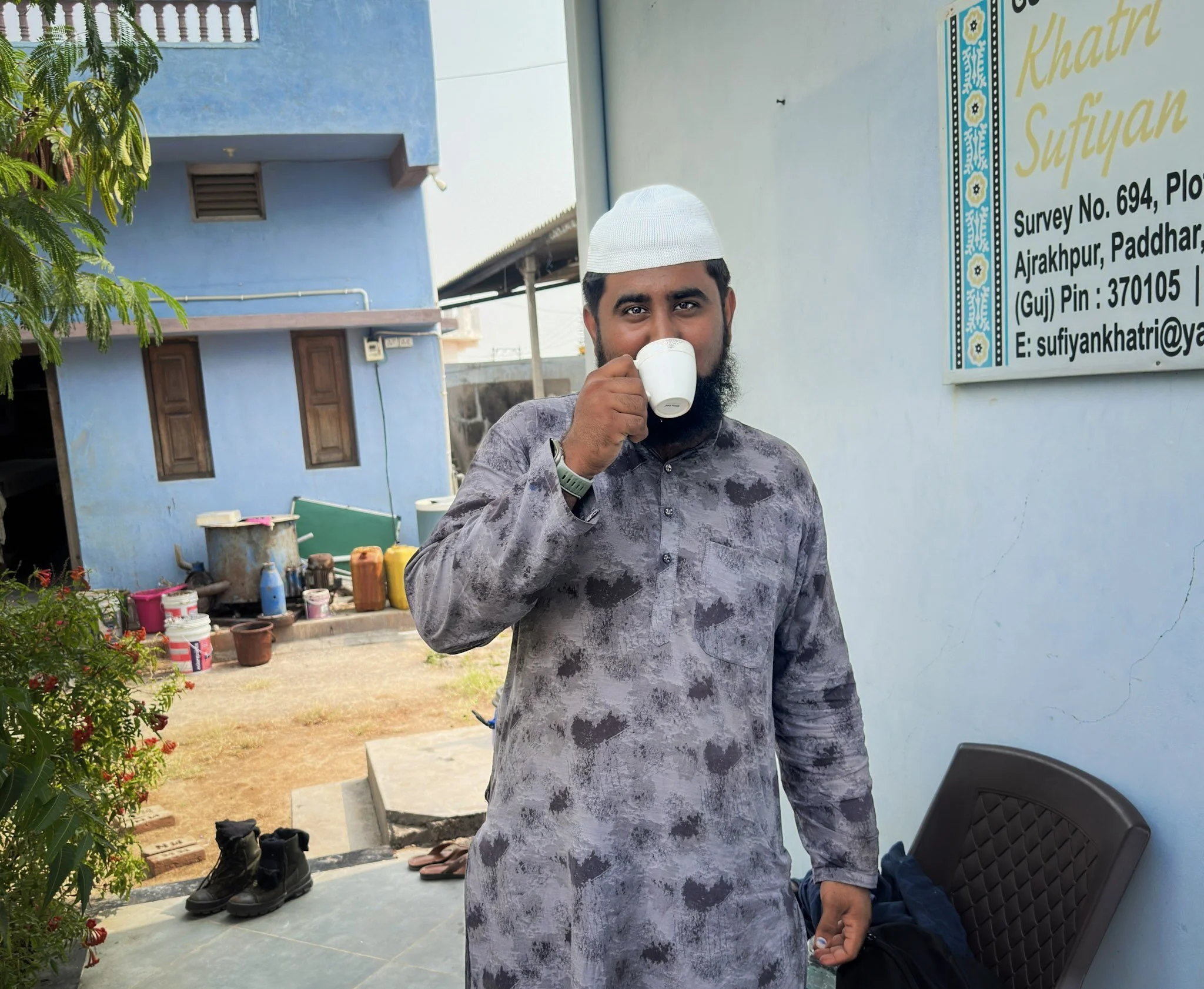Faizan Khatri drinking chai outside Khatri Sufiyan Ismail’s ajrakh workshop in Ajrakhpur — the blue-painted building and the workshop sign visible behind him.