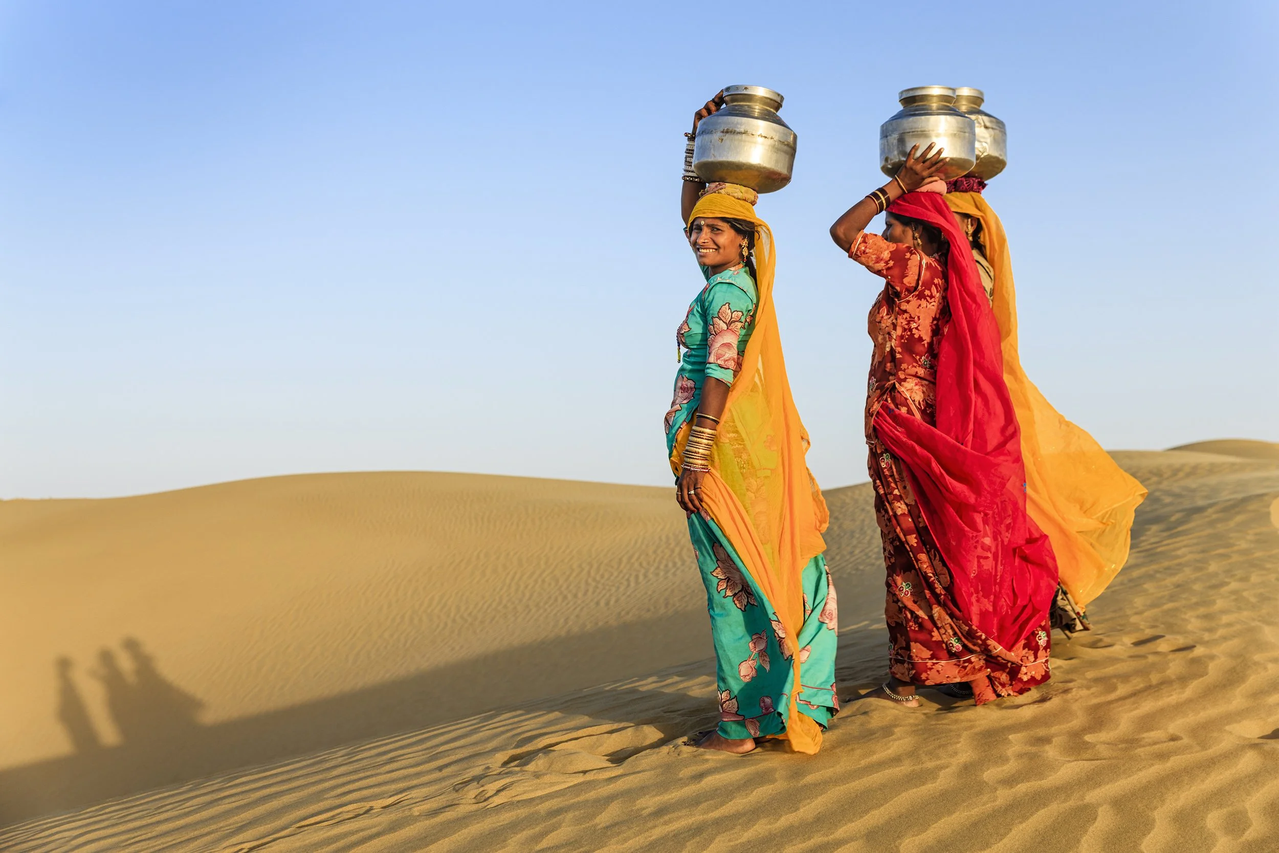 Rajasthani women in vibrant saris carrying water pots on their heads across Thar Desert sand dunes, Kutch.