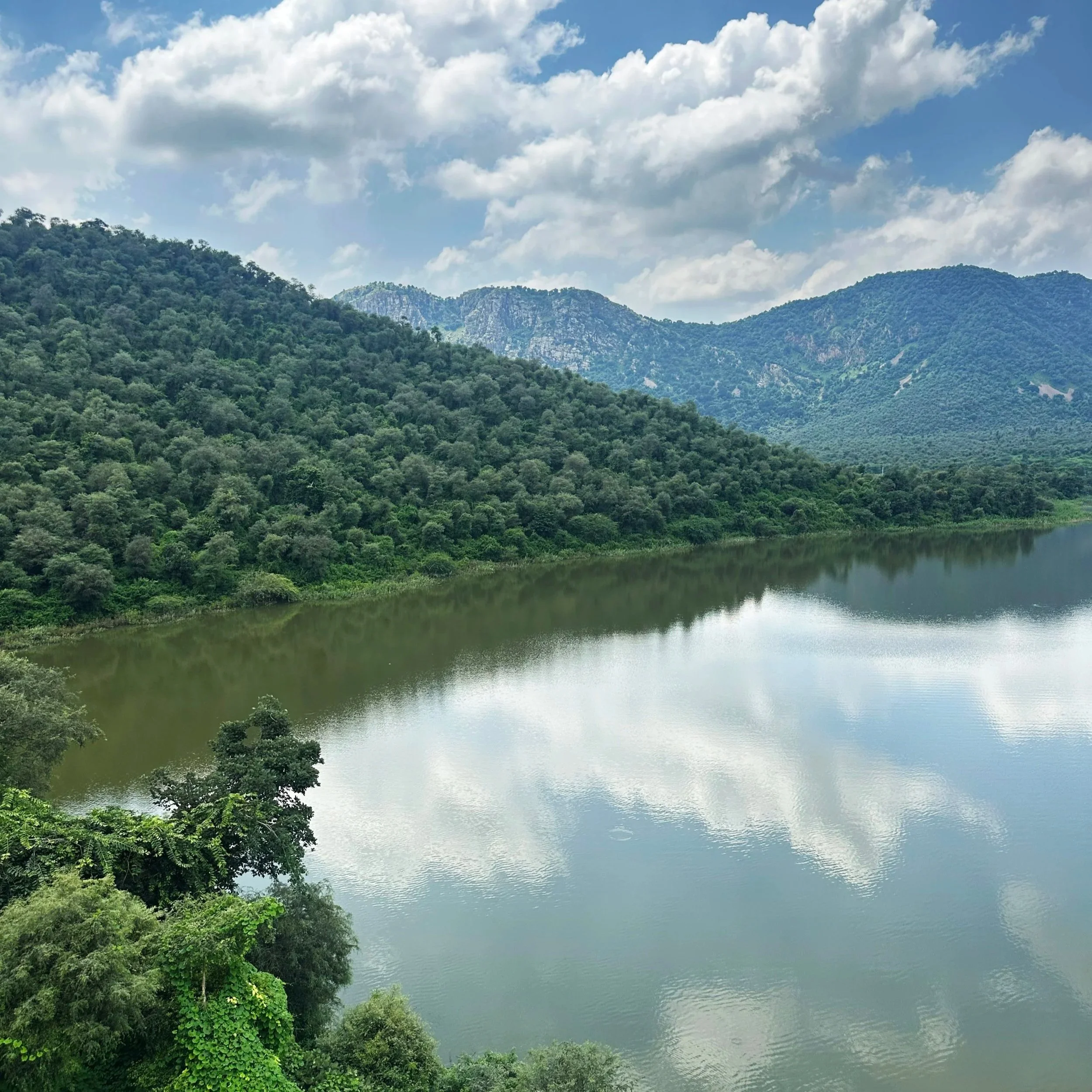 A scenic view of a lake surrounded by green mountains and forest, with partly cloudy skies reflected in the water.