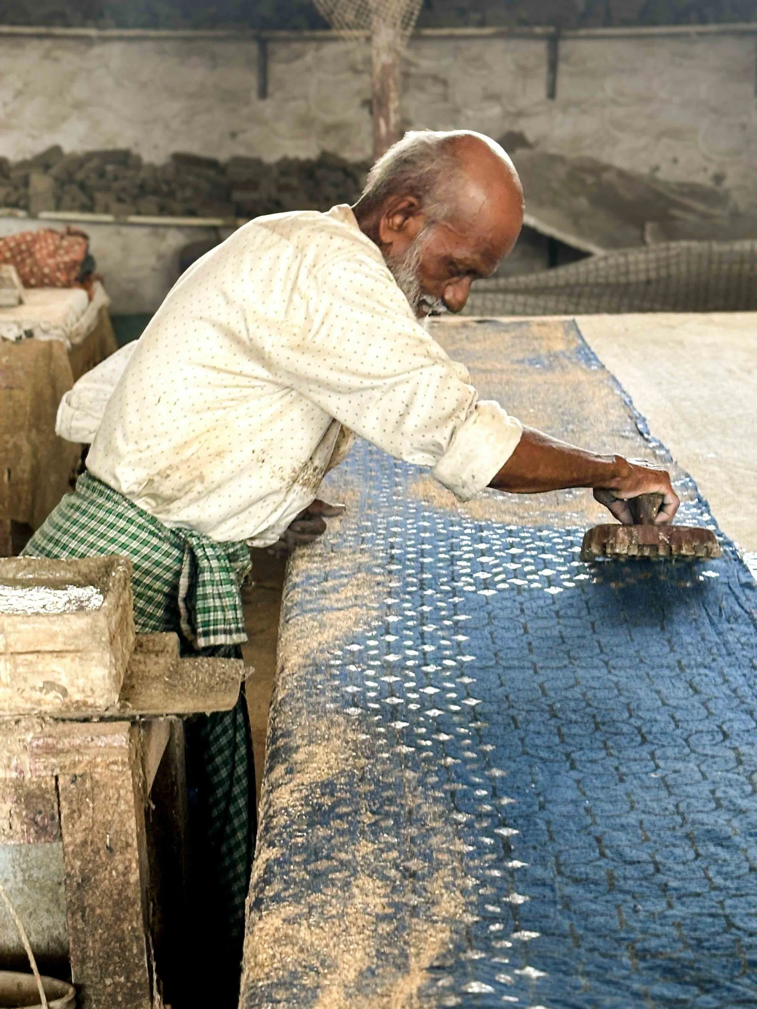 An elderly artisan at a printing table in Pipar, Rajasthan, pressing a carved wooden block onto cotton cloth dusted with sawdust - burada - which binds the mud resist and seals each repeat before dyeing.​​​​​​​​​​​​​​​​