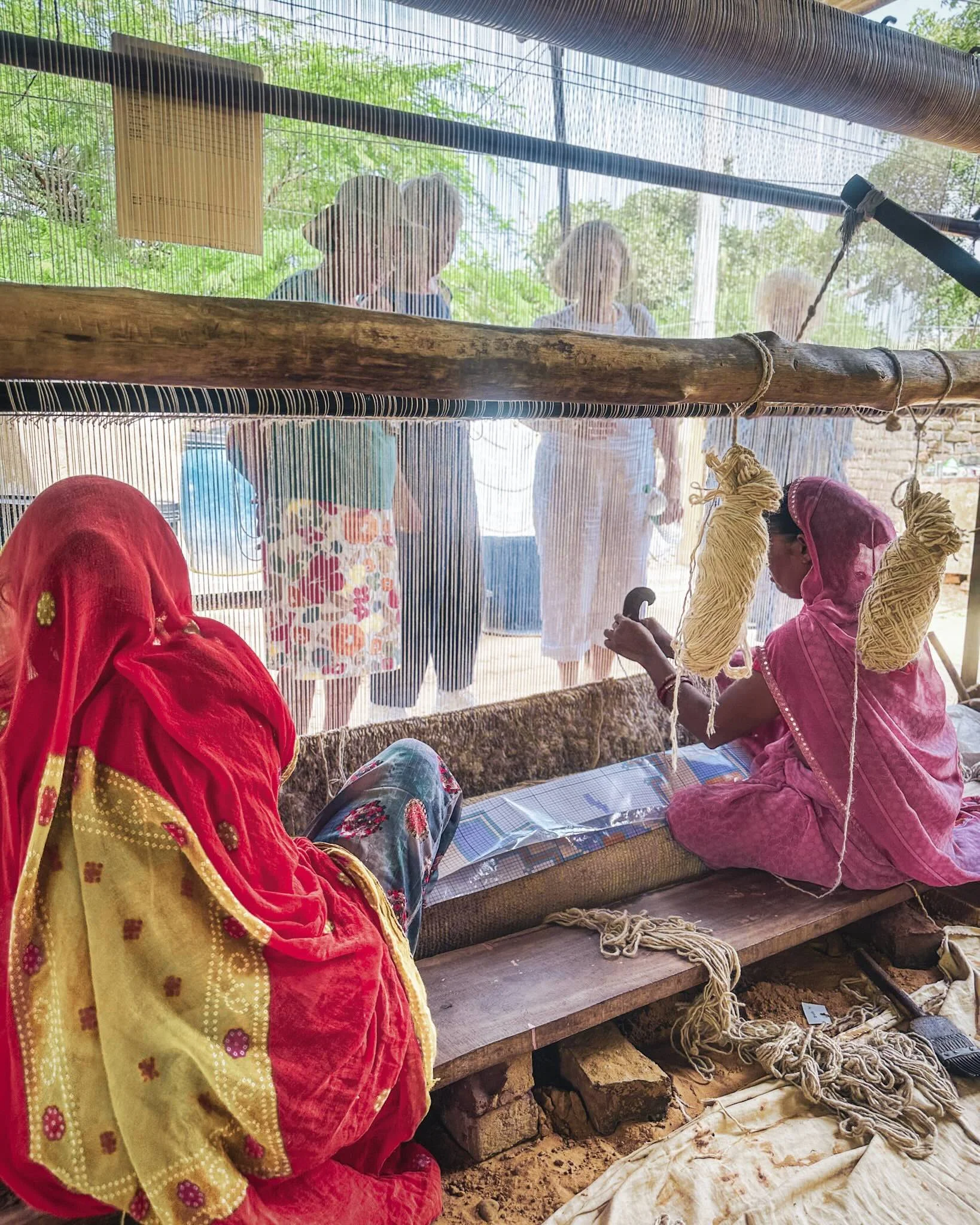 Rajasthani woman weaving at a loom in the foreground, her odhni drawn forward, with a small group of women visitors visible through the warp threads behind her.​​​​​​​​​​​​​​​​
