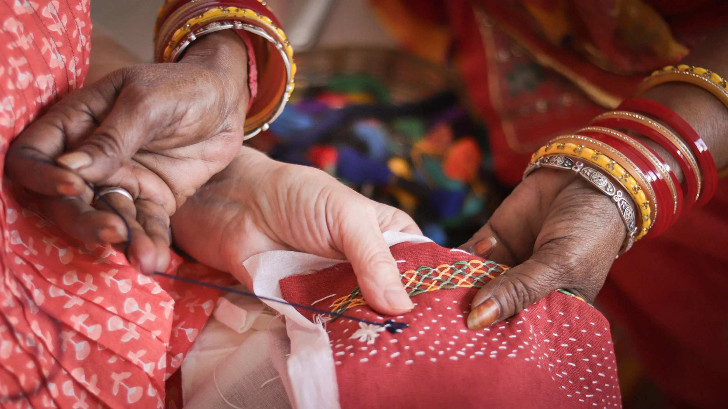 Close-up of two women’s hands, one older and dark-skinned wearing traditional bangles, guiding the other lighter-skinned hand in embroidery on a red fabric with white, green, and yellow thread.