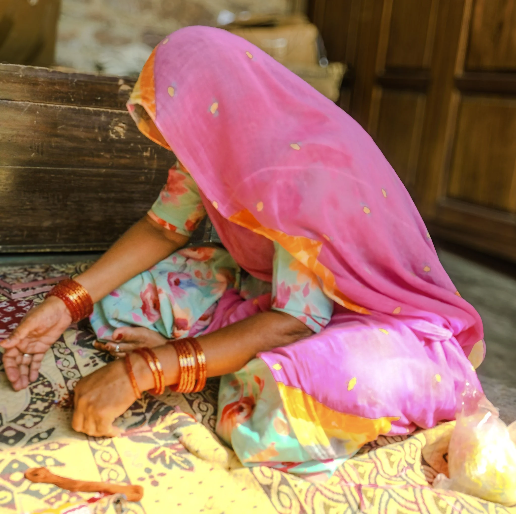 A Rajasthani woman sitting cross-legged and working on the floor at Chandelao, her vivid fuchsia odhni falling forward over her head and shoulders, wearing red lac bangles and a pale floral ghagra.​​​​​​​​​​​​​​​​