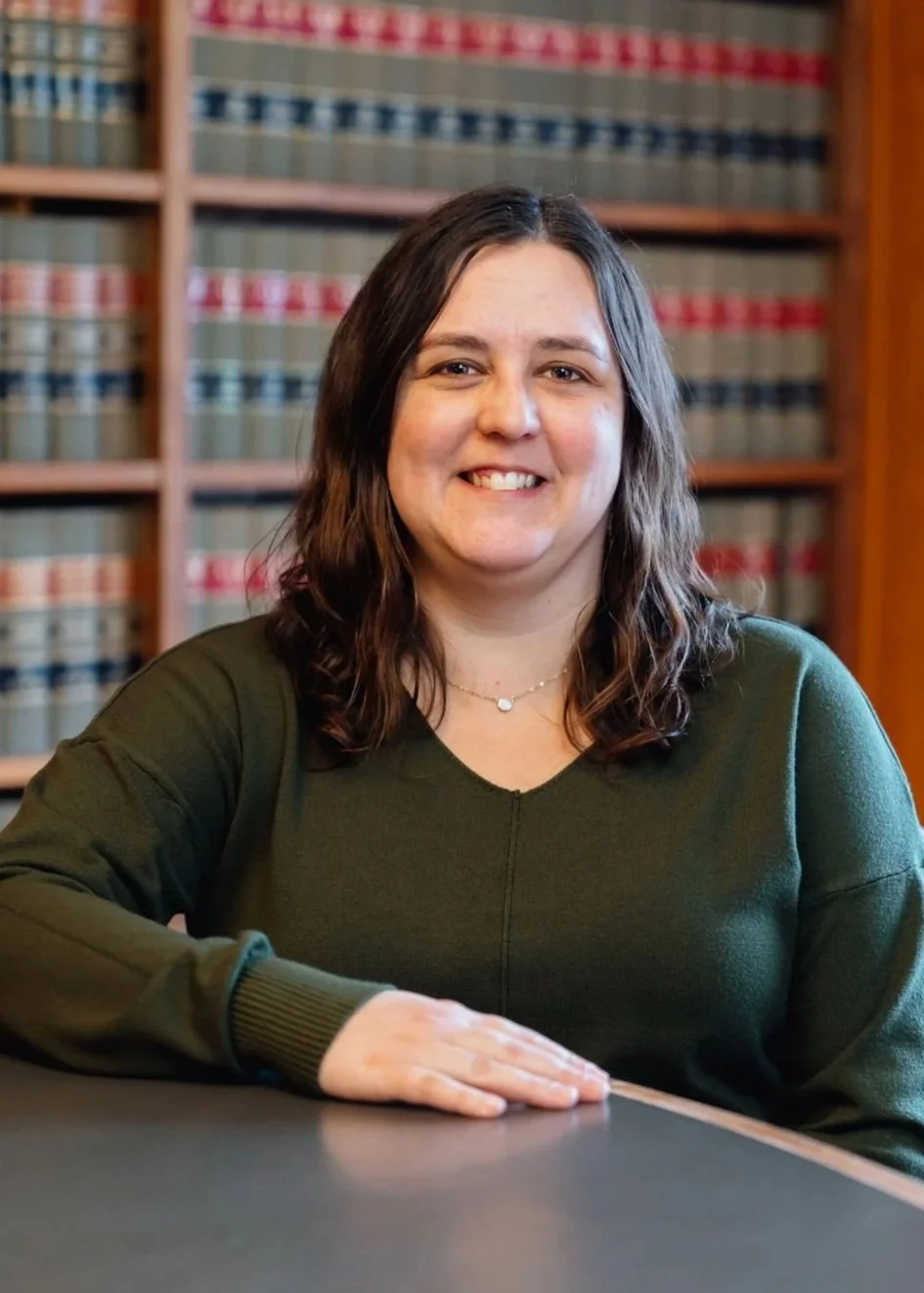 A woman with shoulder-length dark hair sitting at a table in a library, with bookshelves filled with law books in the background.