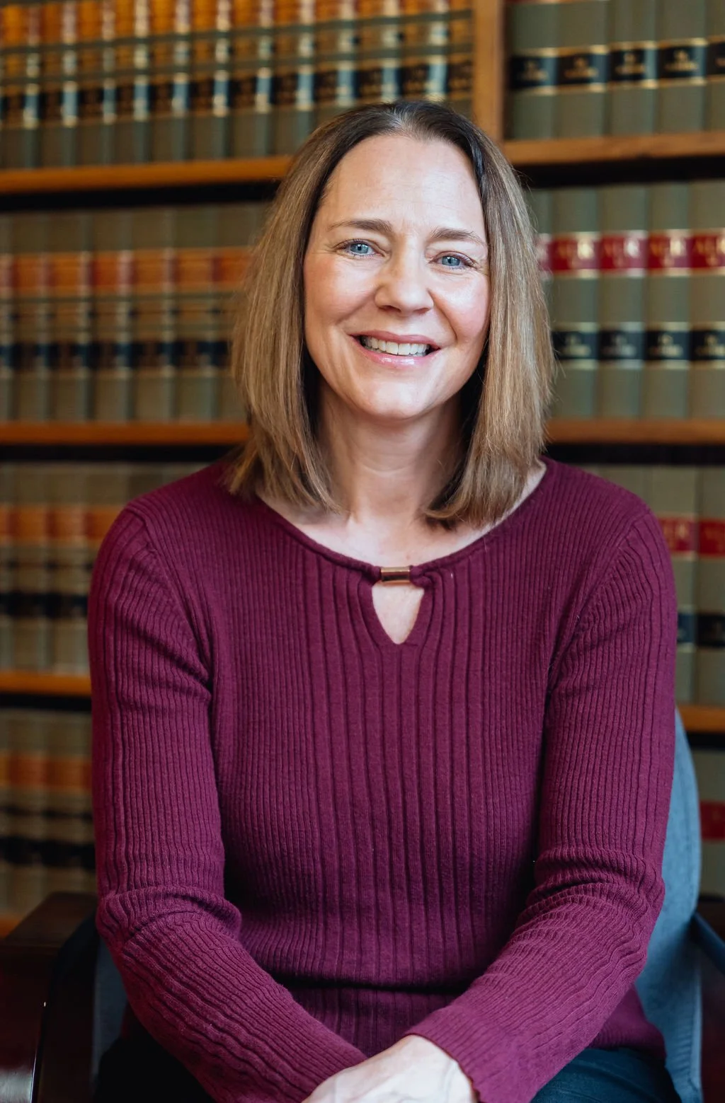 A smiling woman with shoulder-length brown hair and blue eyes, wearing a maroon ribbed sweater, sitting in front of a bookshelf filled with law books.