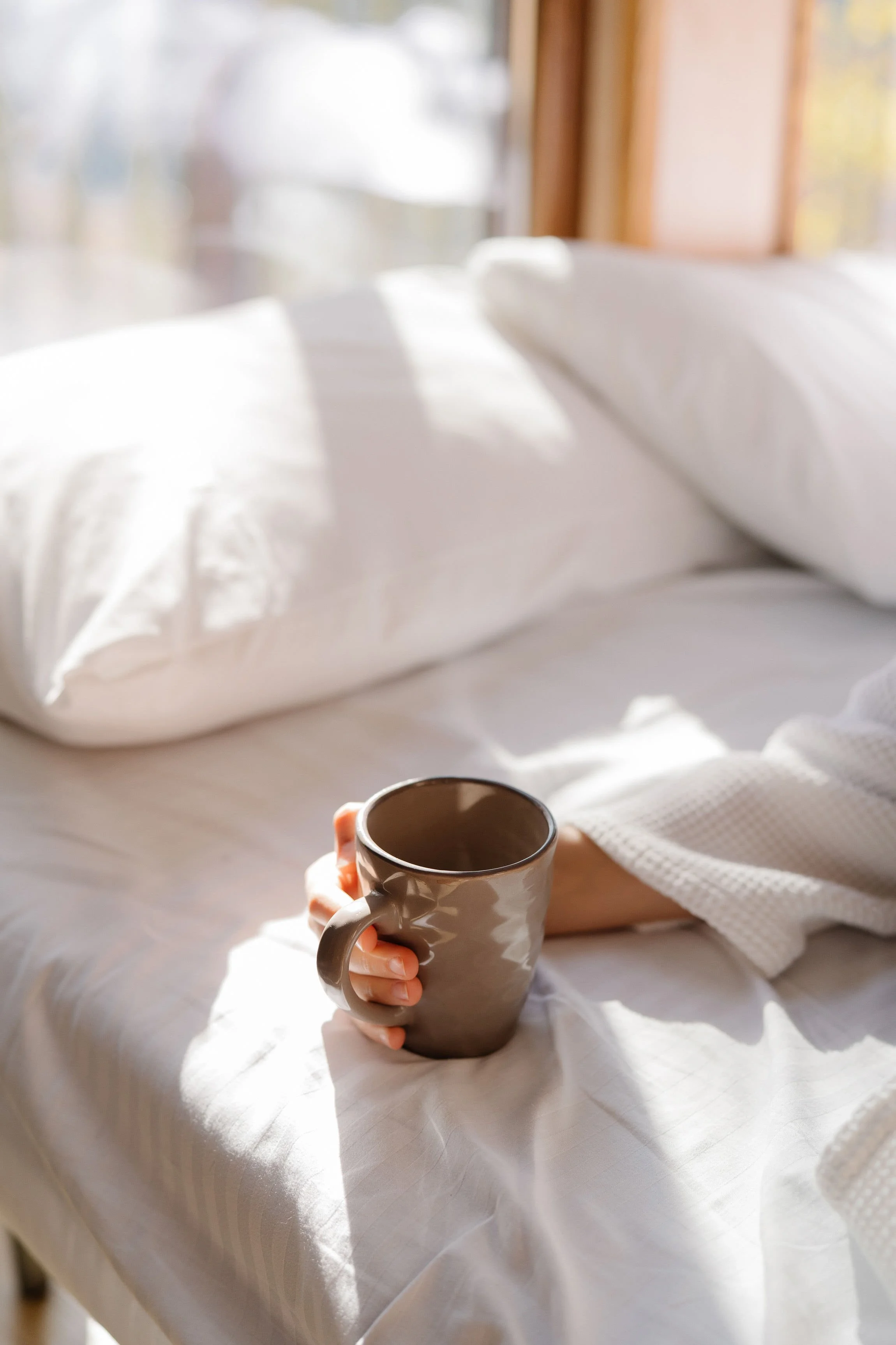 A person sitting in bed holding a brown mug, with sunlight streaming through a window, white bedding, and wood window frames visible in the background.