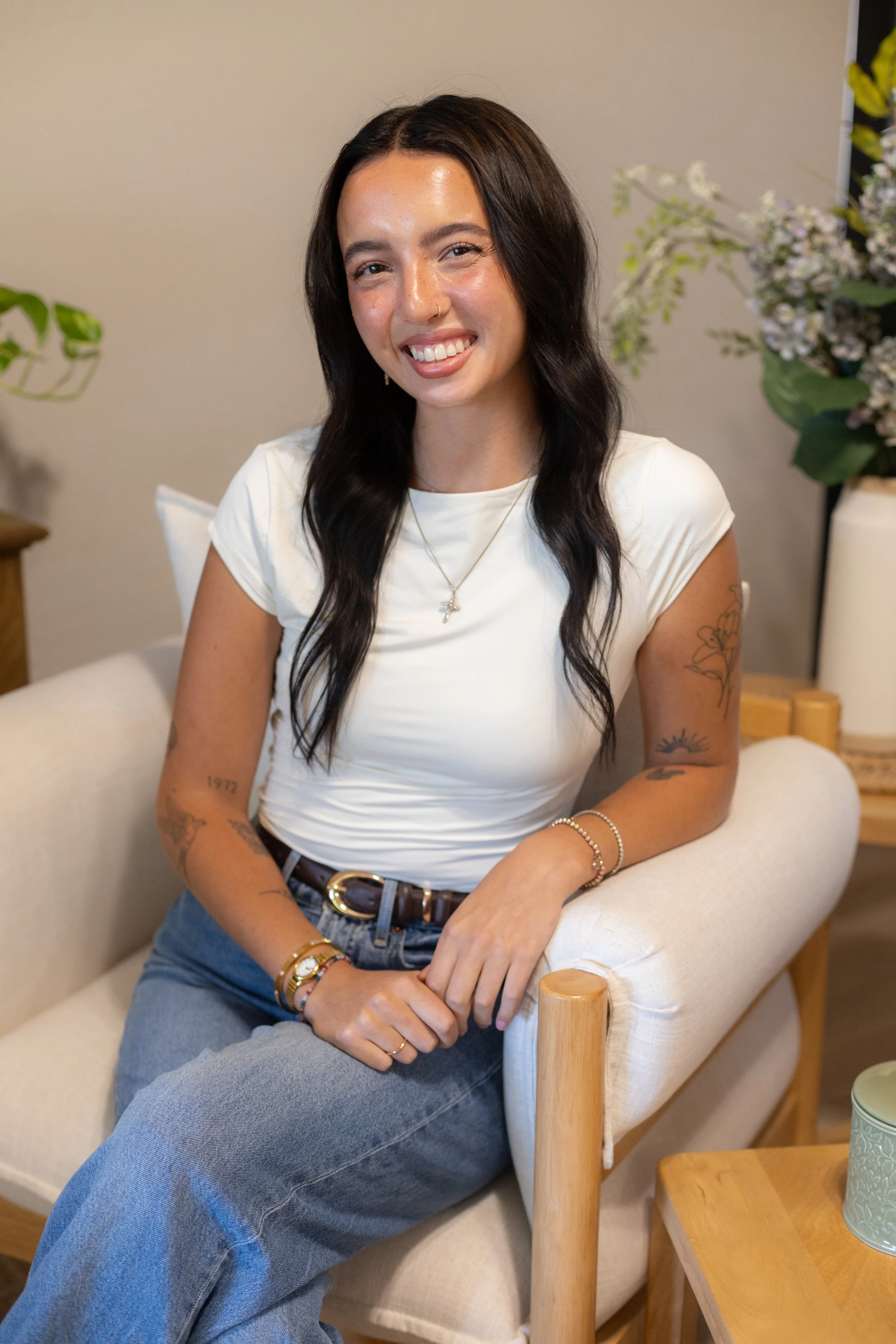 A young woman with long dark hair, tattoos on her arms, and a nose ring, sitting on a cream-colored armchair in a cozy room with plants and flowers in the background. She is smiling and wearing a white t-shirt and blue jeans.