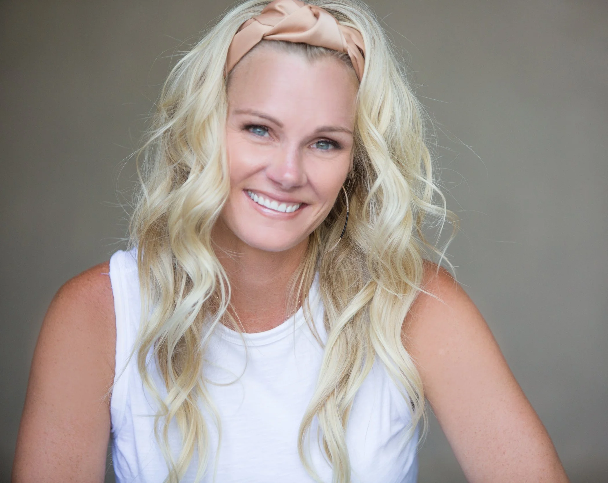 A young woman with long, wavy blonde hair smiling and wearing a light pink headband and a white sleeveless top.