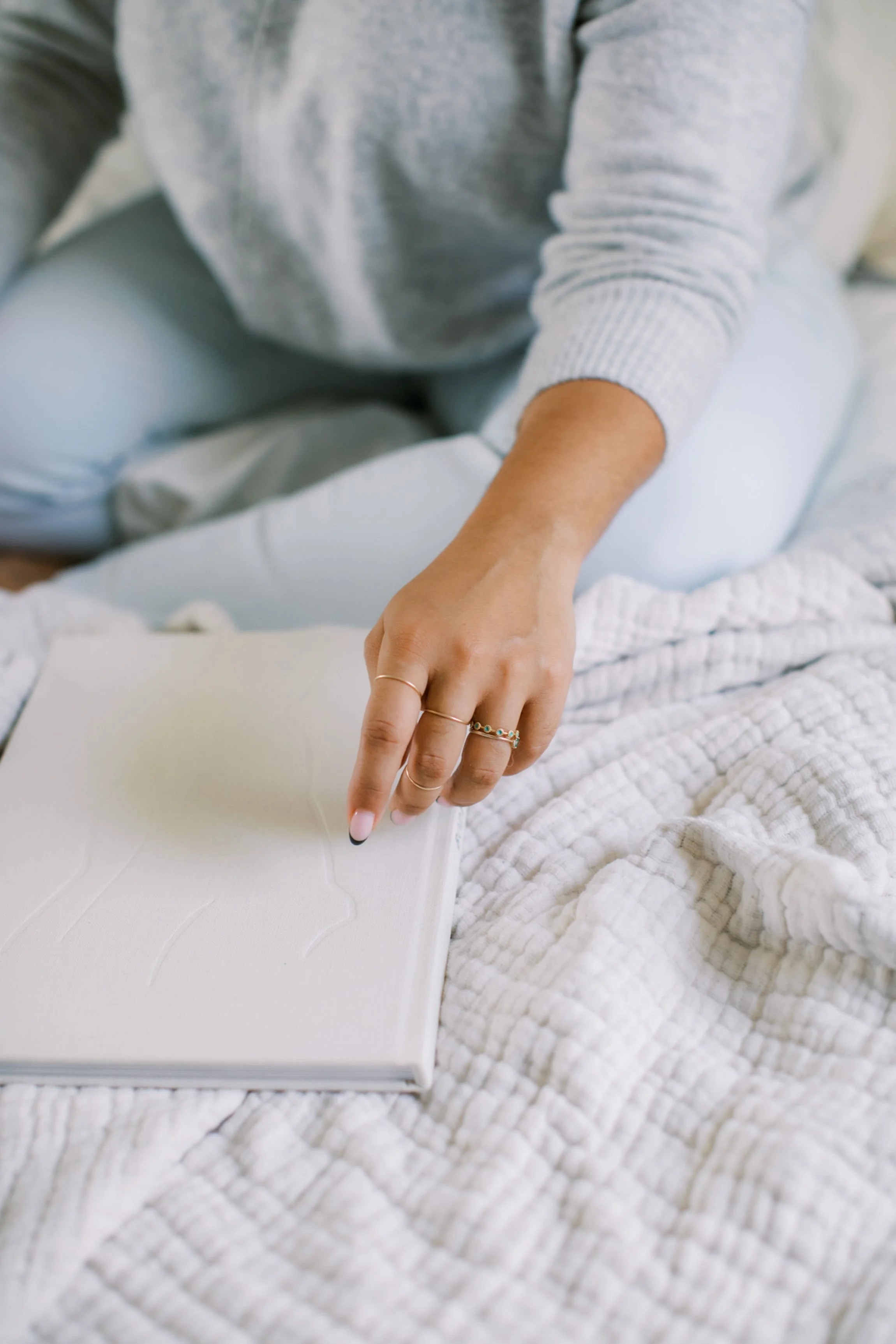 Close-up of a person's hand pointing at a white, textured book or journal on a bed with a cream-colored blanket, wearing multiple rings on fingers, dressed in a gray long-sleeve shirt.