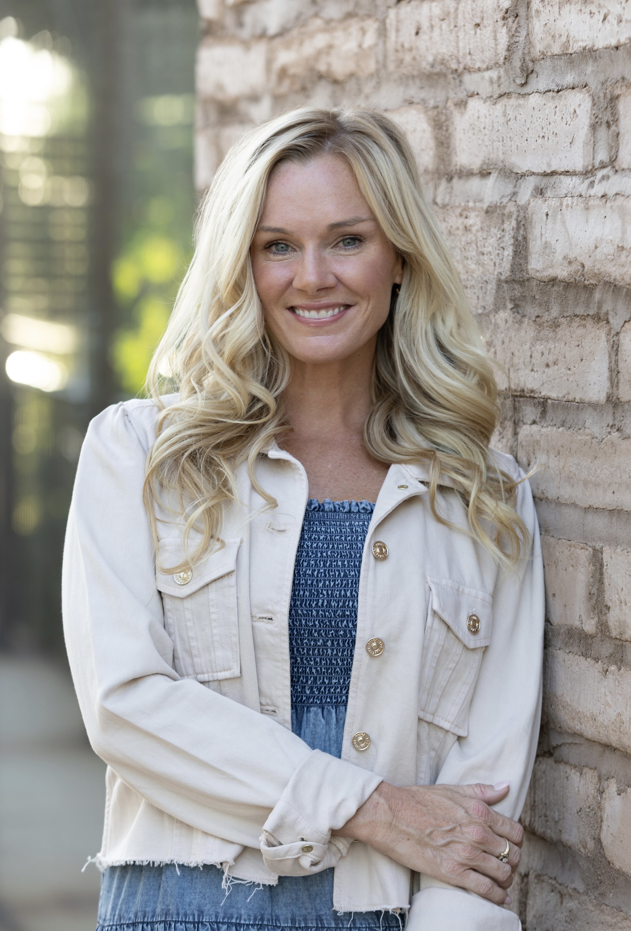 A woman with long blonde hair, wearing a light-colored jacket, standing beside a brick wall. She is smiling and has her arms crossed.