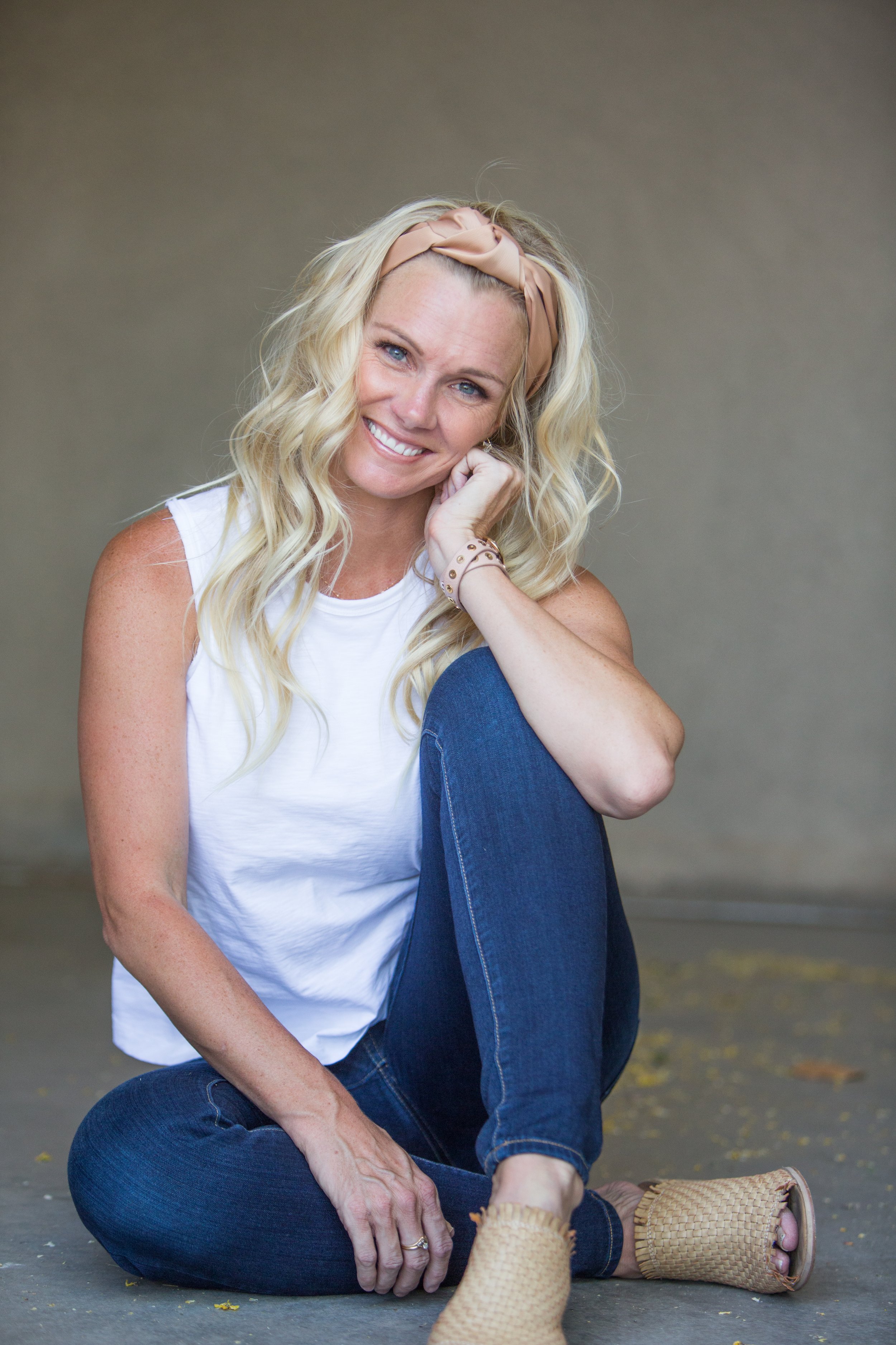 A woman with blonde hair wearing a beige headband, white sleeveless top, and blue jeans sitting on the ground and smiling at the camera, with a child's foot in the foreground wearing tan woven sandals.