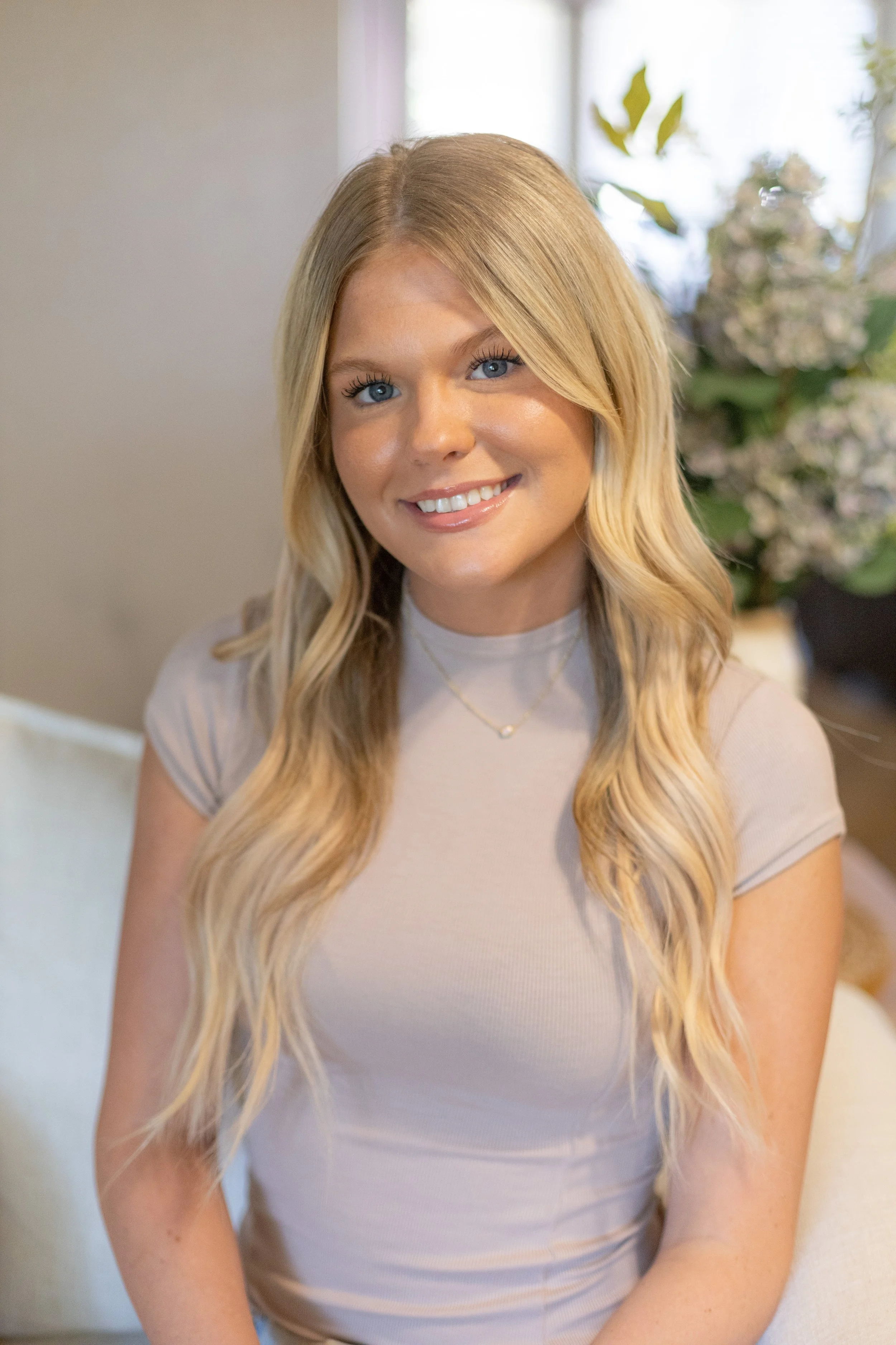 A woman with long, wavy blonde hair and blue eyes smiling and wearing a light-colored t-shirt and a delicate necklace, sitting indoors near a window with plants and flowers in the background.