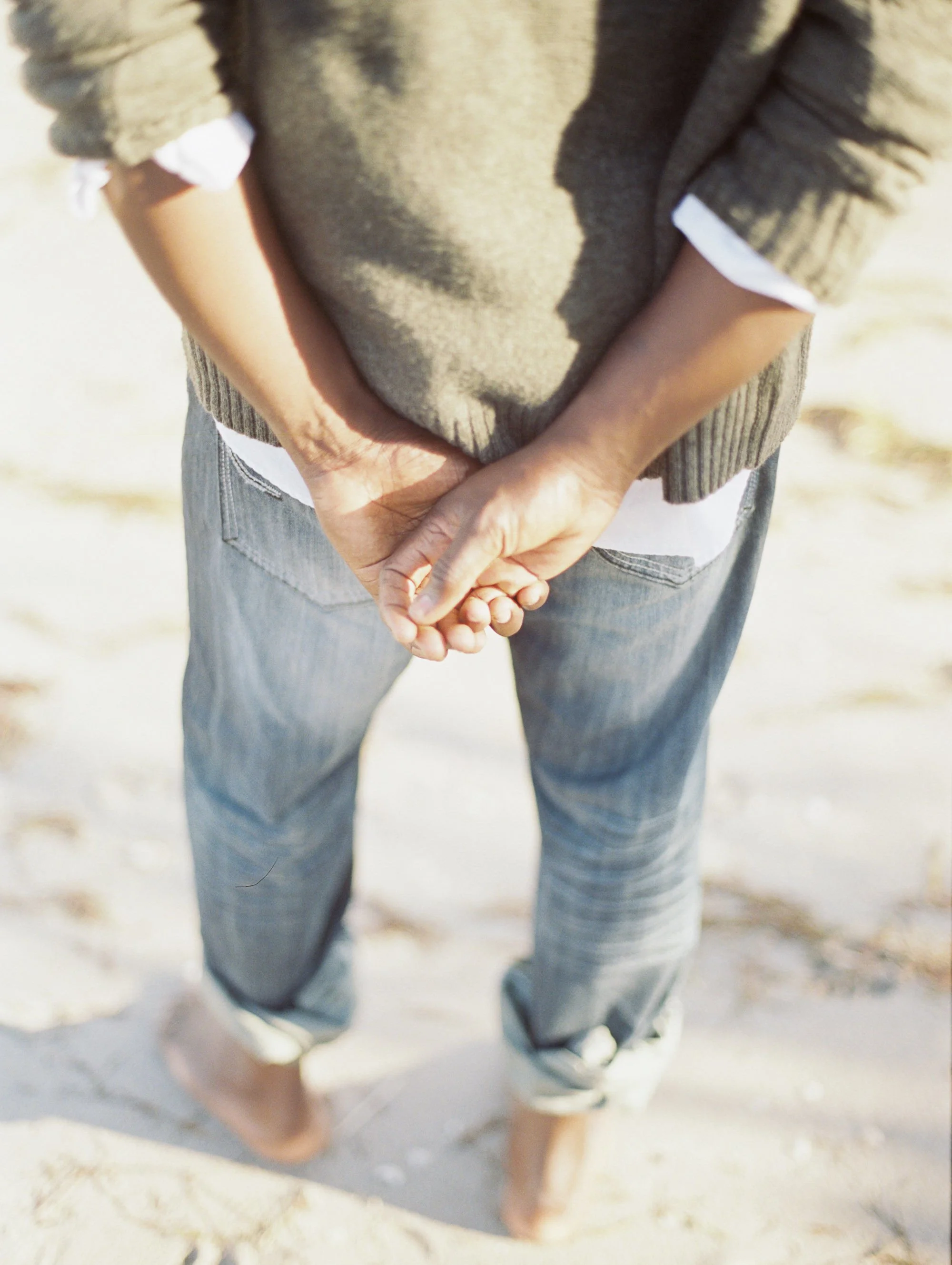 Person holding hands behind their back, wearing a brown sweater over a white shirt and jeans, standing on sandy ground.