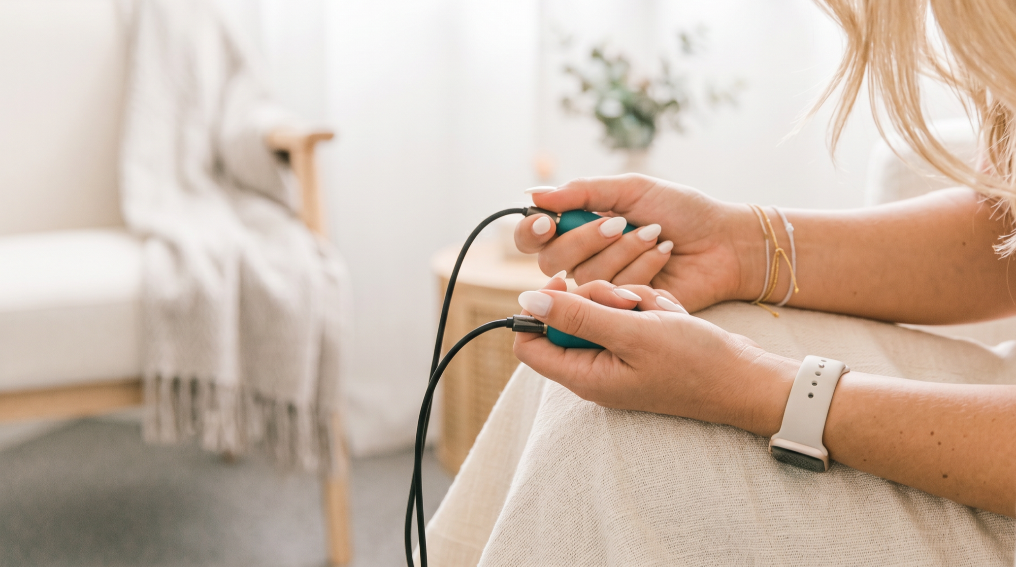 Person lying on a bed or couch, holding a handheld pulse oximeter on their finger, wearing a smartwatch on their wrist. In the background, there's a person in pajamas standing near a bed with a striped blanket.